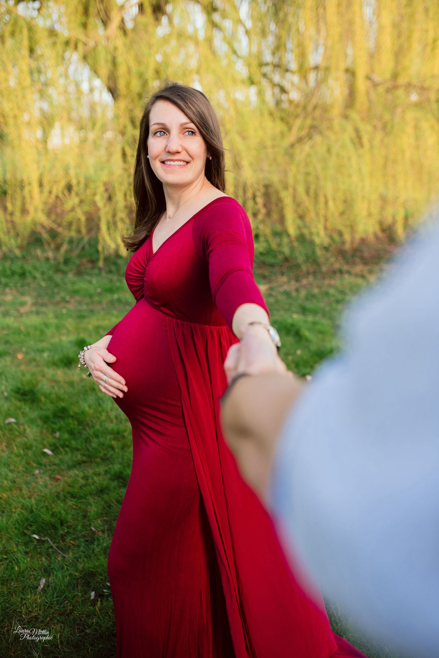 A maternity photo of a woman in a flowing red dress outdoors with autumn foliage.