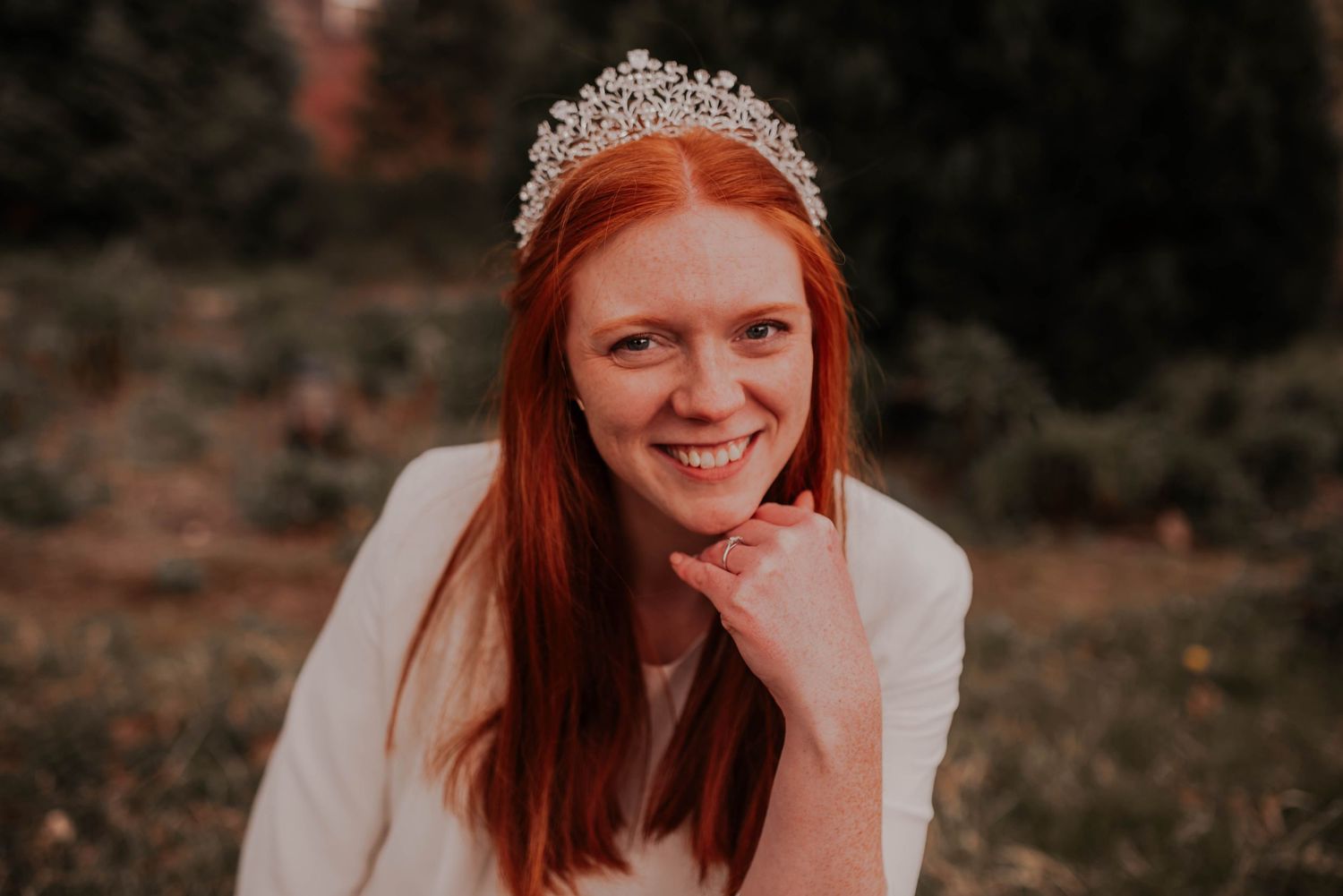 A person in white wearing a crystal tiara poses outdoors with a gentle smile.
