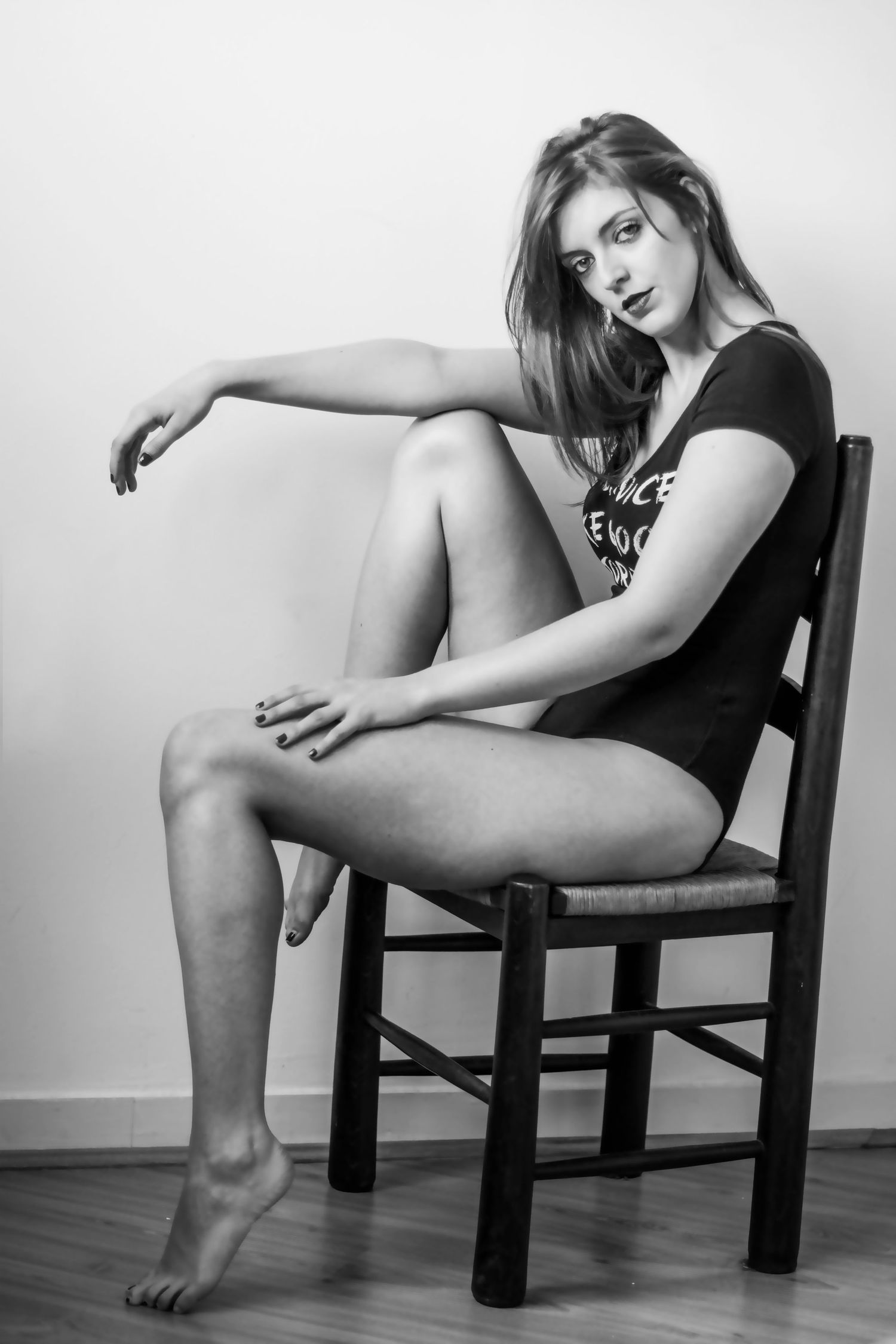 Black and white portrait of model posing on wooden chair in casual attire.