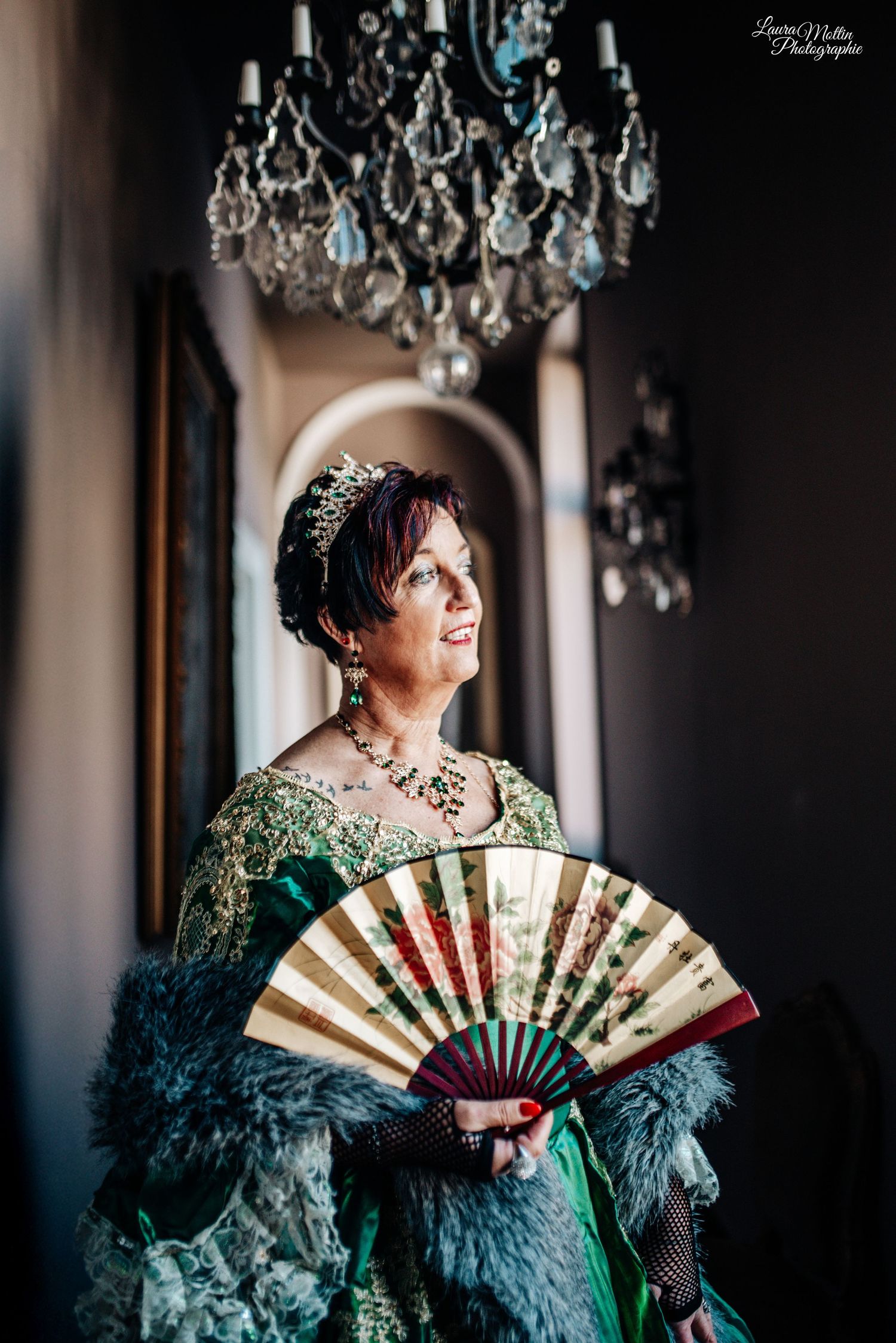 Theatrical portrait with ornate fan and emerald green costume under crystal chandelier lighting.