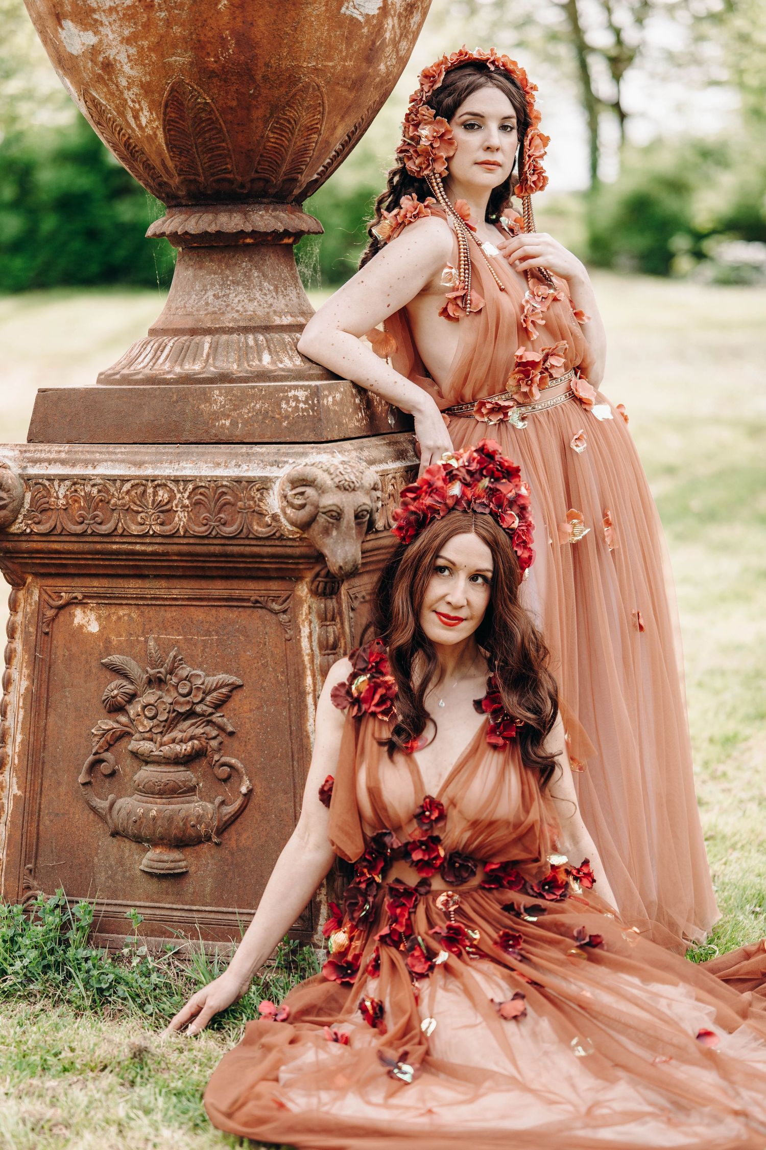 Two women in floral rust-colored gowns with flower crowns pose elegantly by an ornate garden urn.
