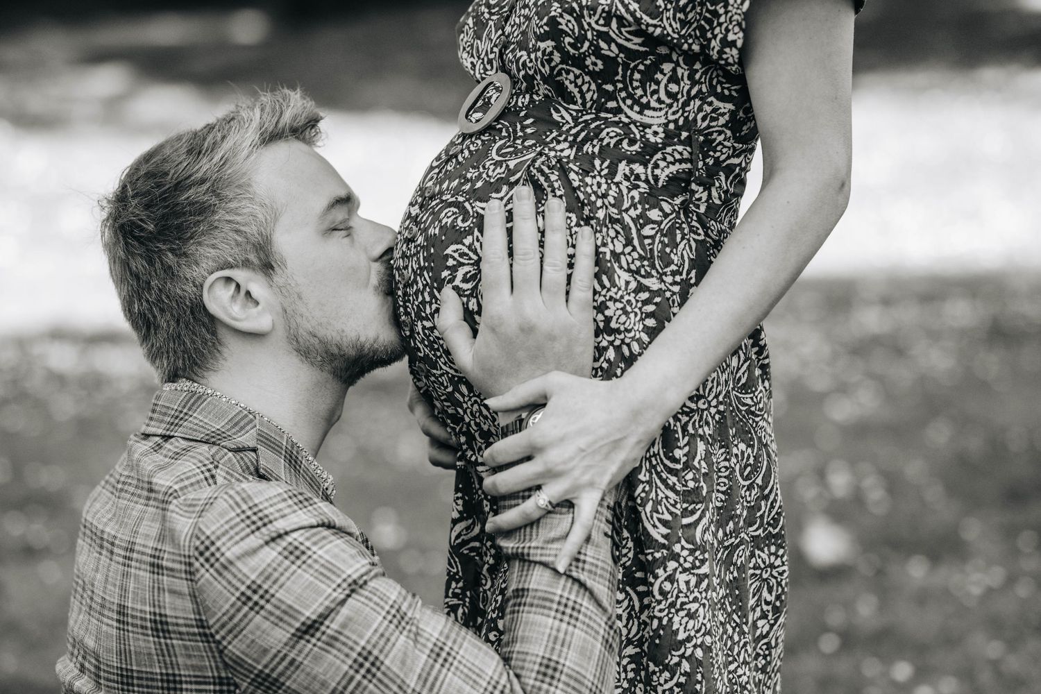 Black and white photo of a man kissing a pregnant woman's belly while holding it gently outdoors.