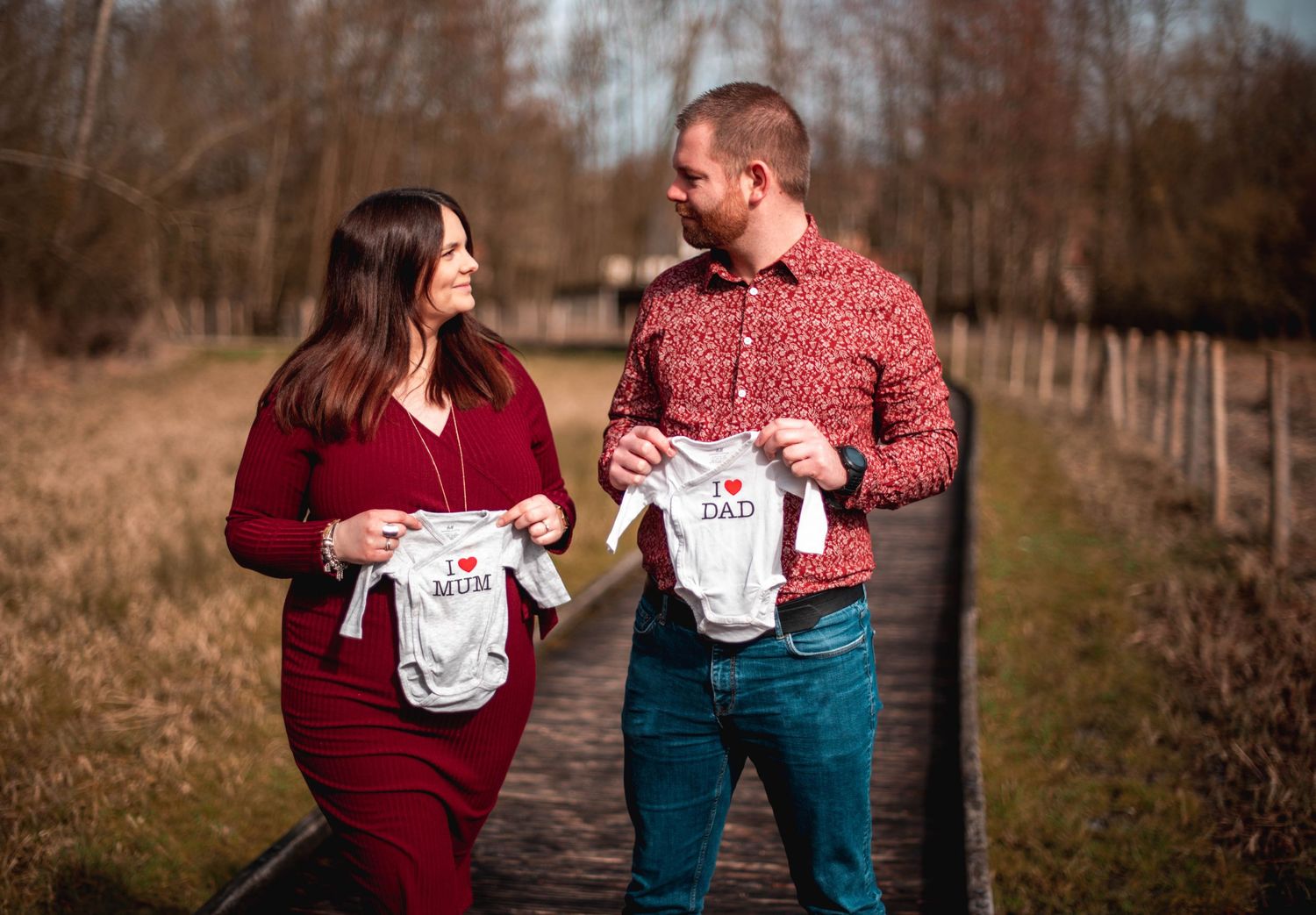 Autumn maternity photoshoot with couple holding baby clothes in woodland setting.