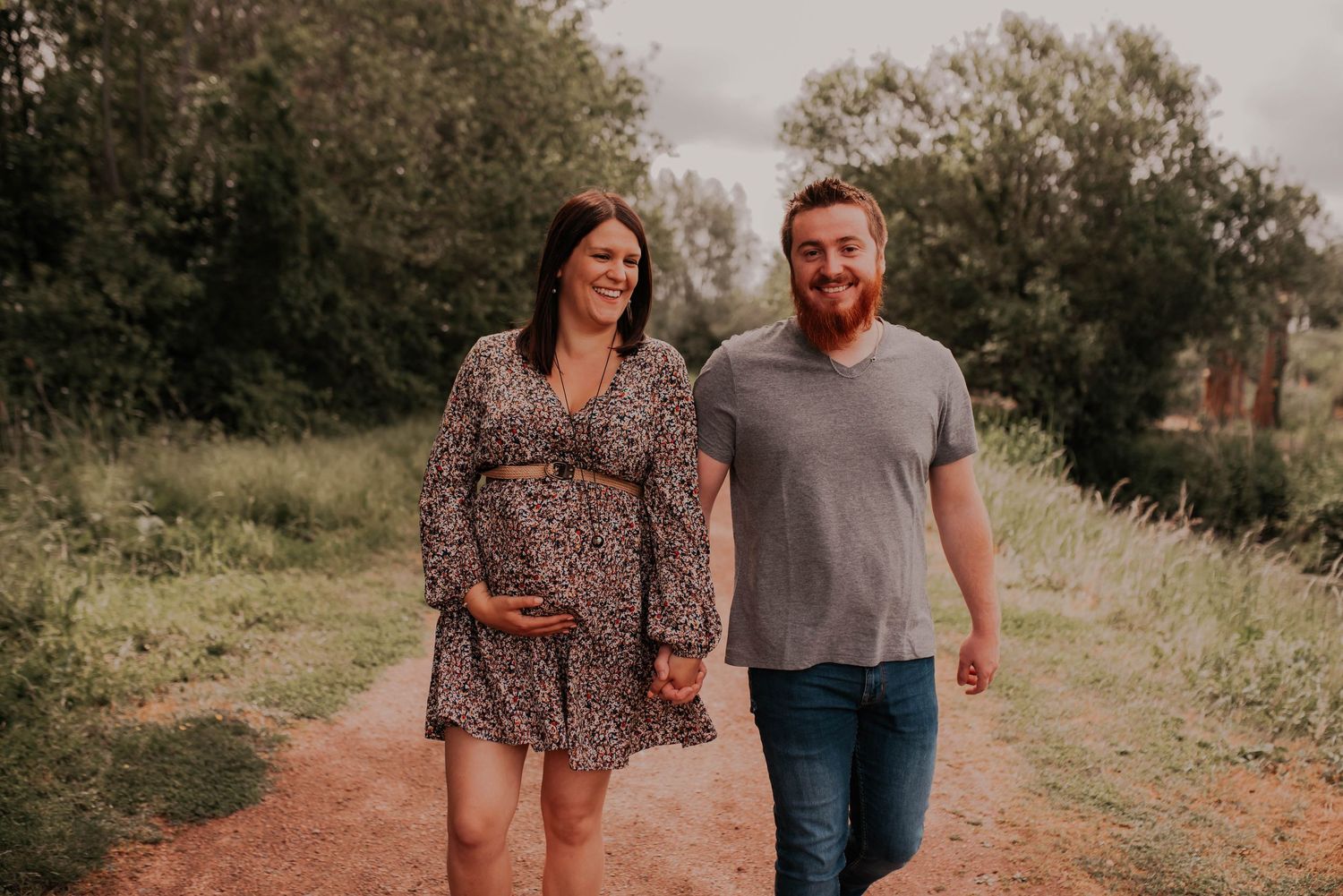 Expecting couple walking together on rustic trail surrounded by greenery.