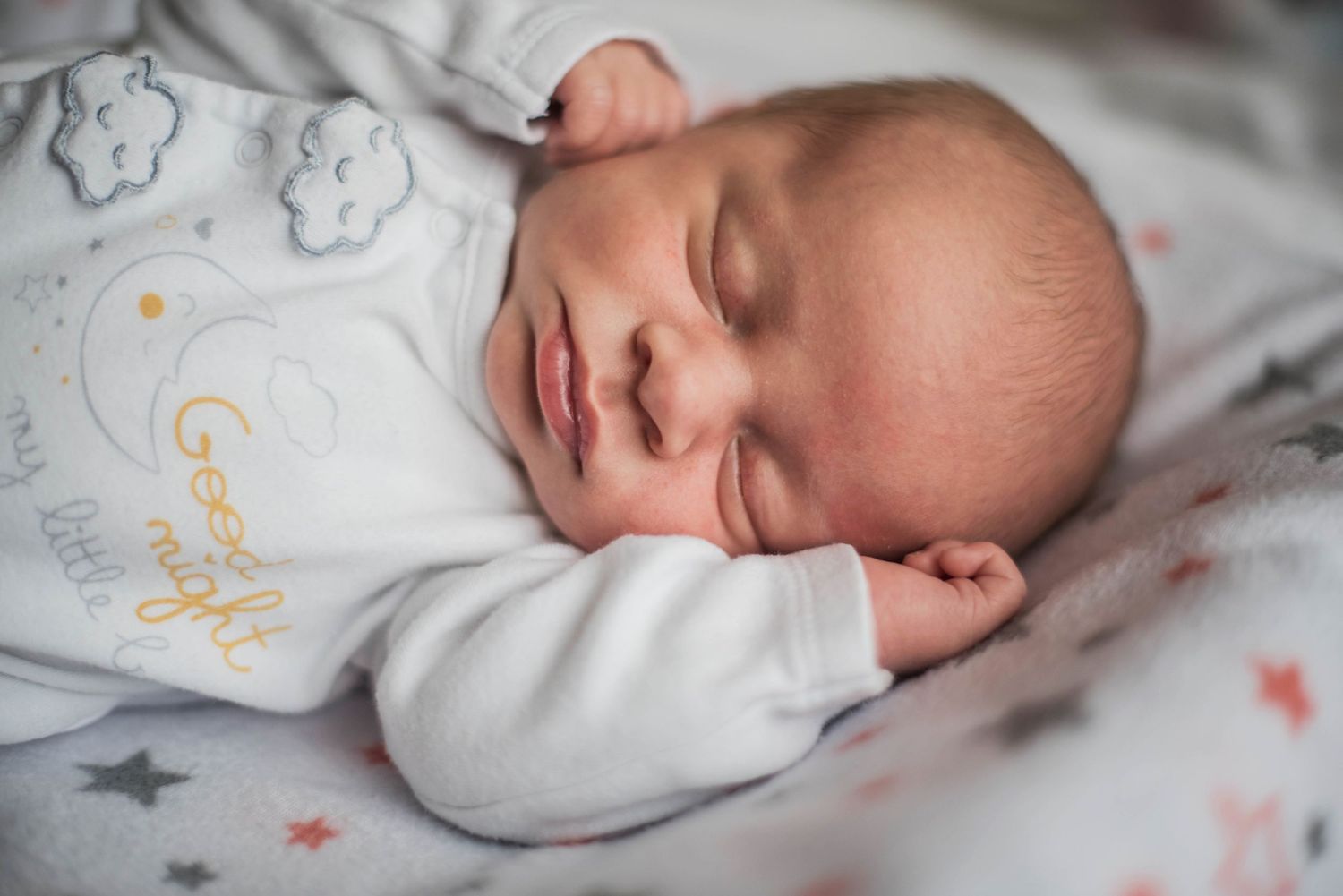 Sleeping newborn wearing star-patterned white outfit captured in gentle natural light.