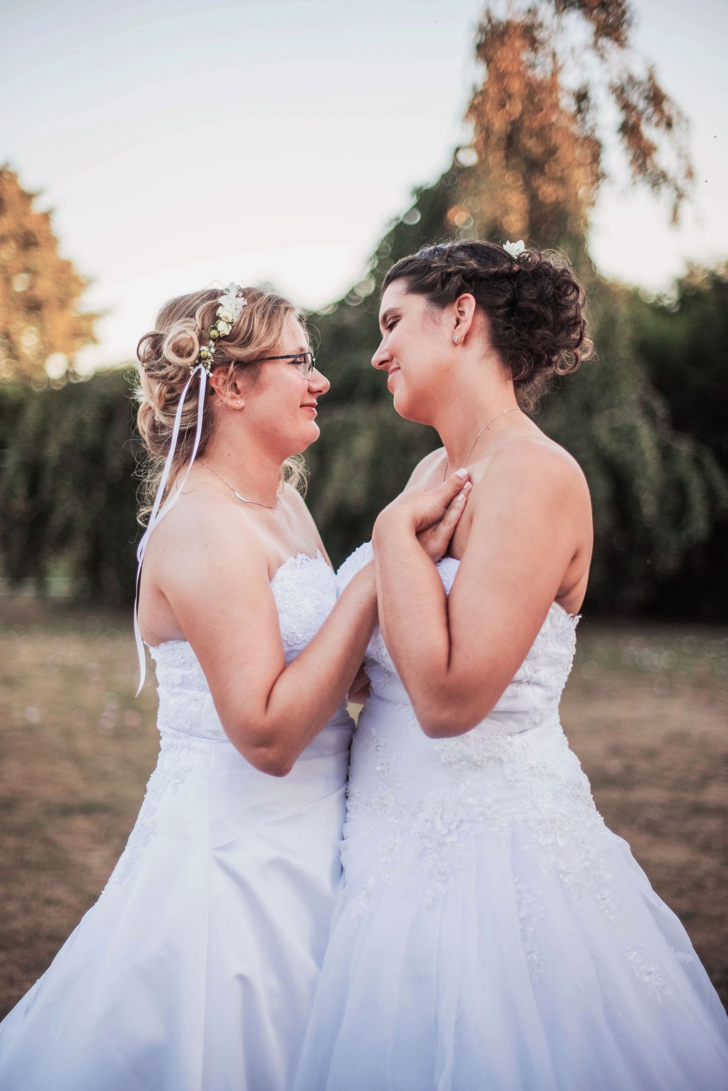 Two brides in white wedding dresses share an intimate moment at sunset in a natural outdoor setting.