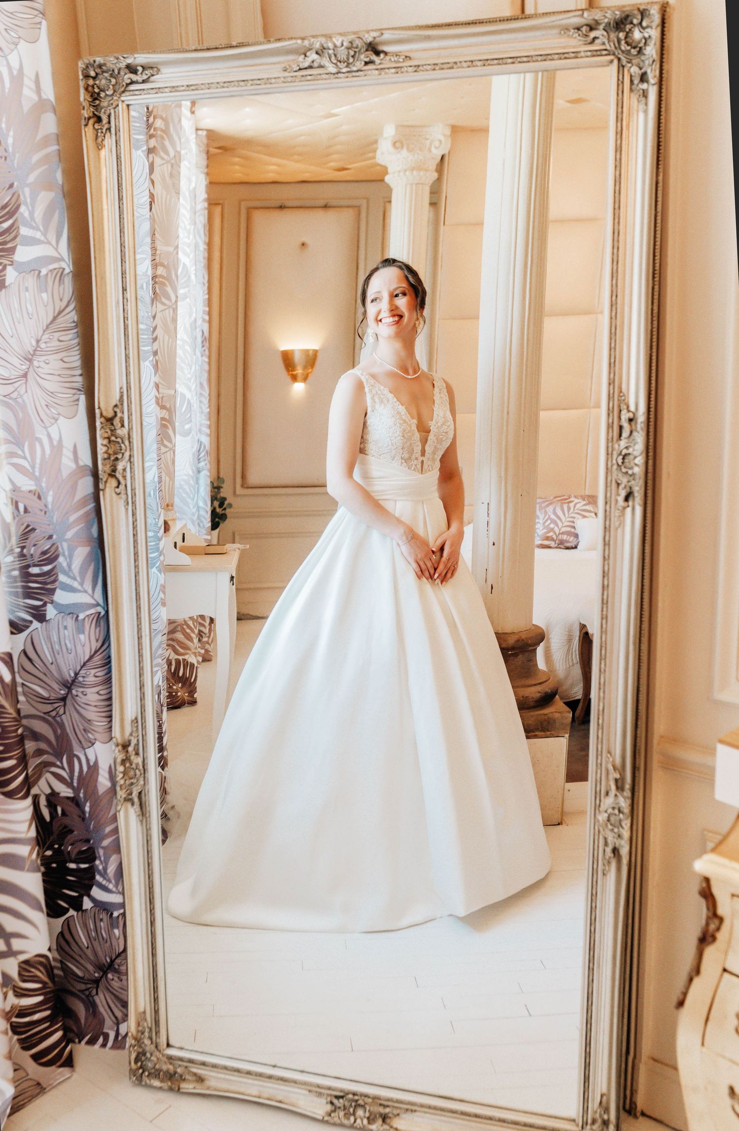 Smiling bride in elegant white ball gown with lace bodice reflected in ornate silver mirror in a luxurious bridal suite.