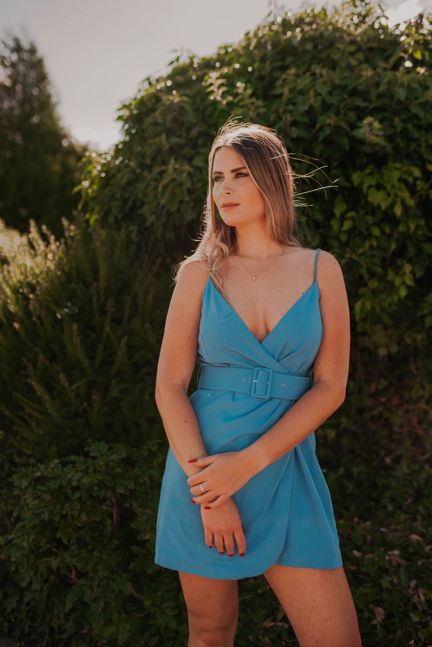 Woman in a bright blue summer dress poses against greenery during golden hour.