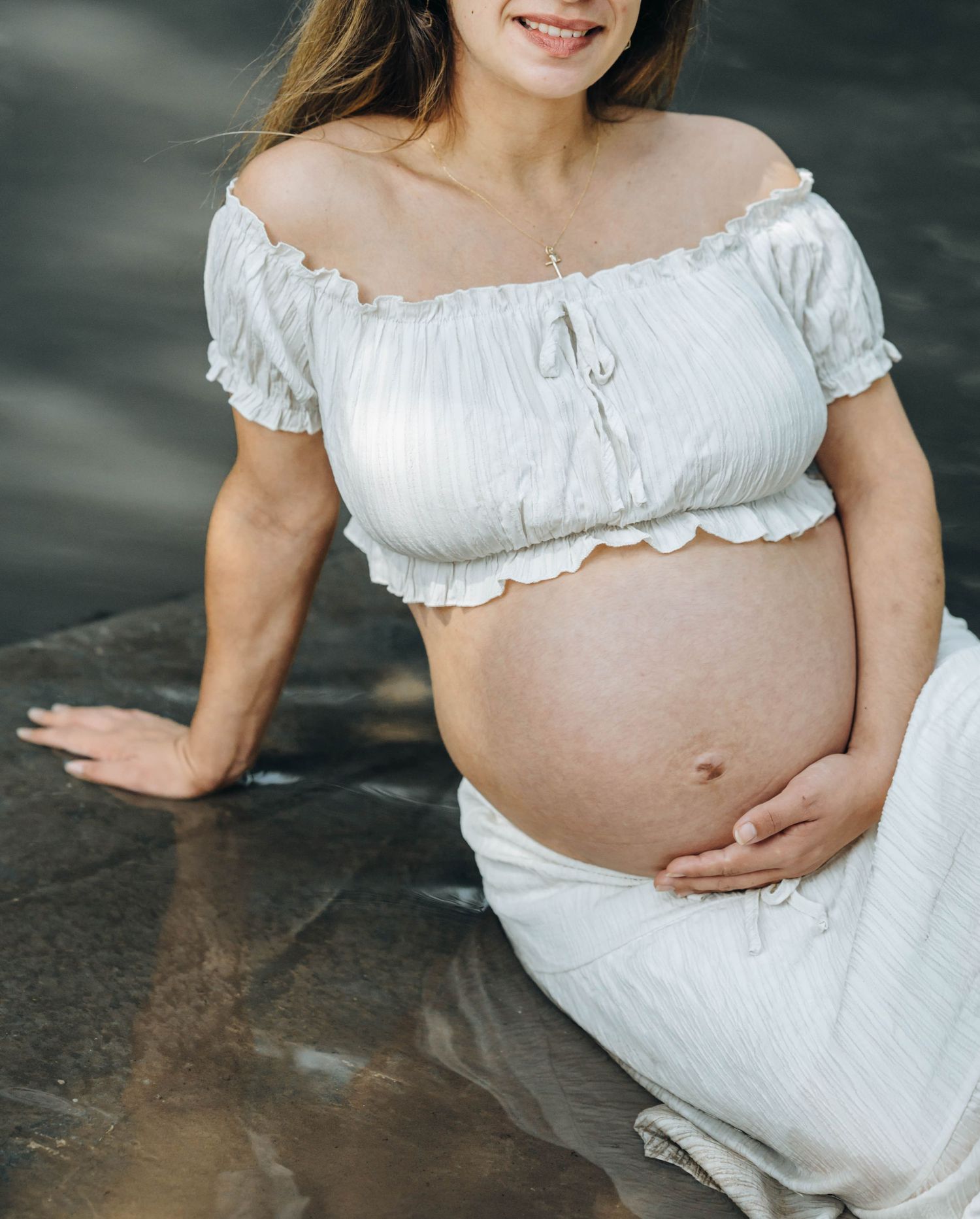 Smiling pregnant woman in white off-shoulder crop top and skirt cradling her baby bump outdoors.