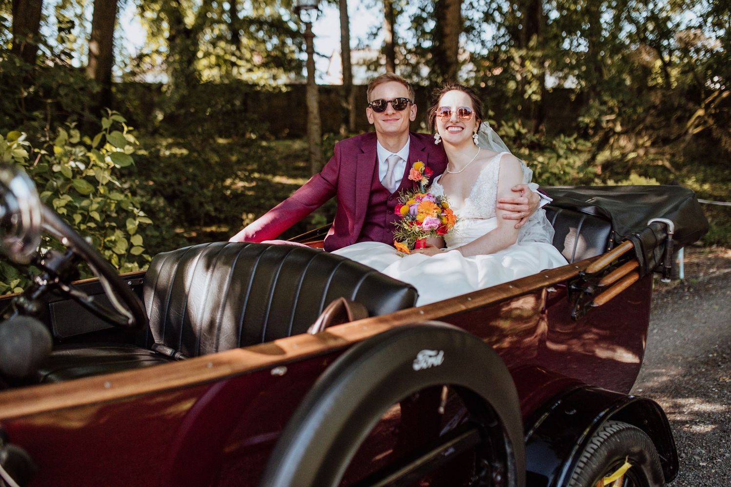 Bride and groom in sunglasses riding in a vintage burgundy convertible car on their wedding day.