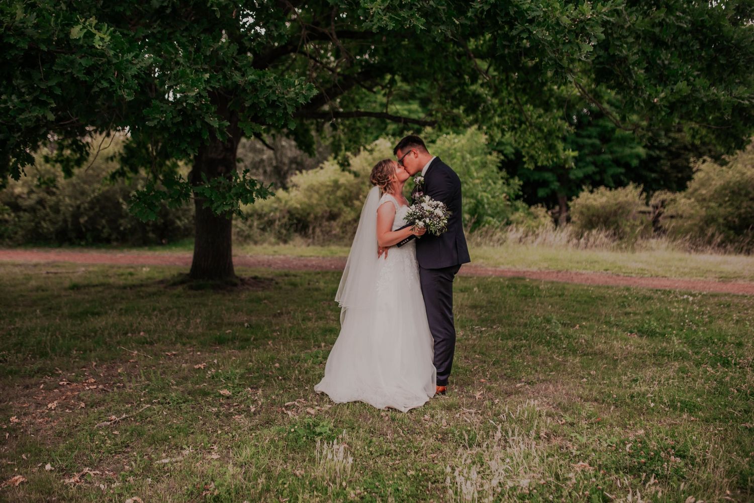 A couple embraces under a large tree during their outdoor wedding photoshoot.