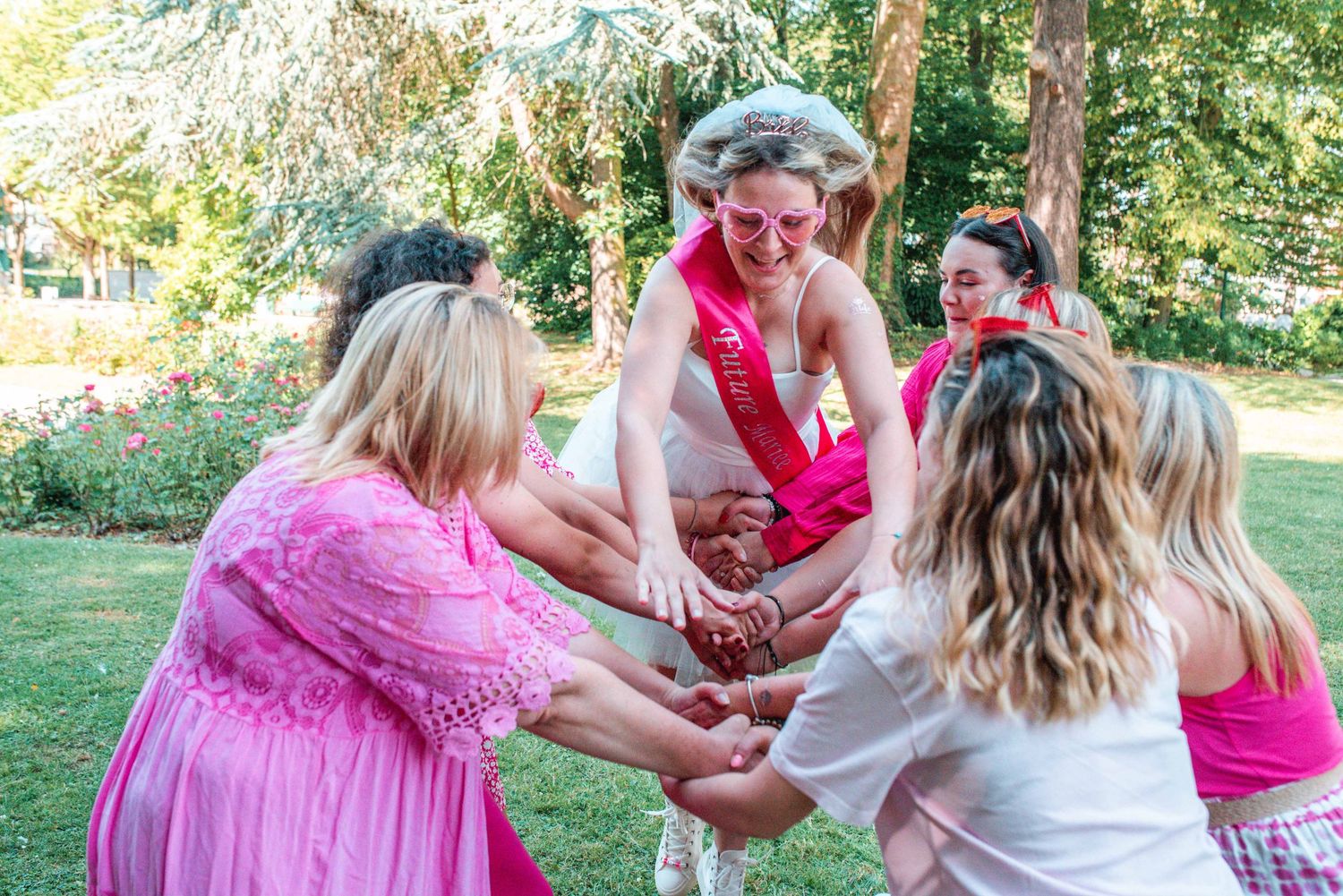 A joyful group plays party games outdoors in pink dresses and casual attire.