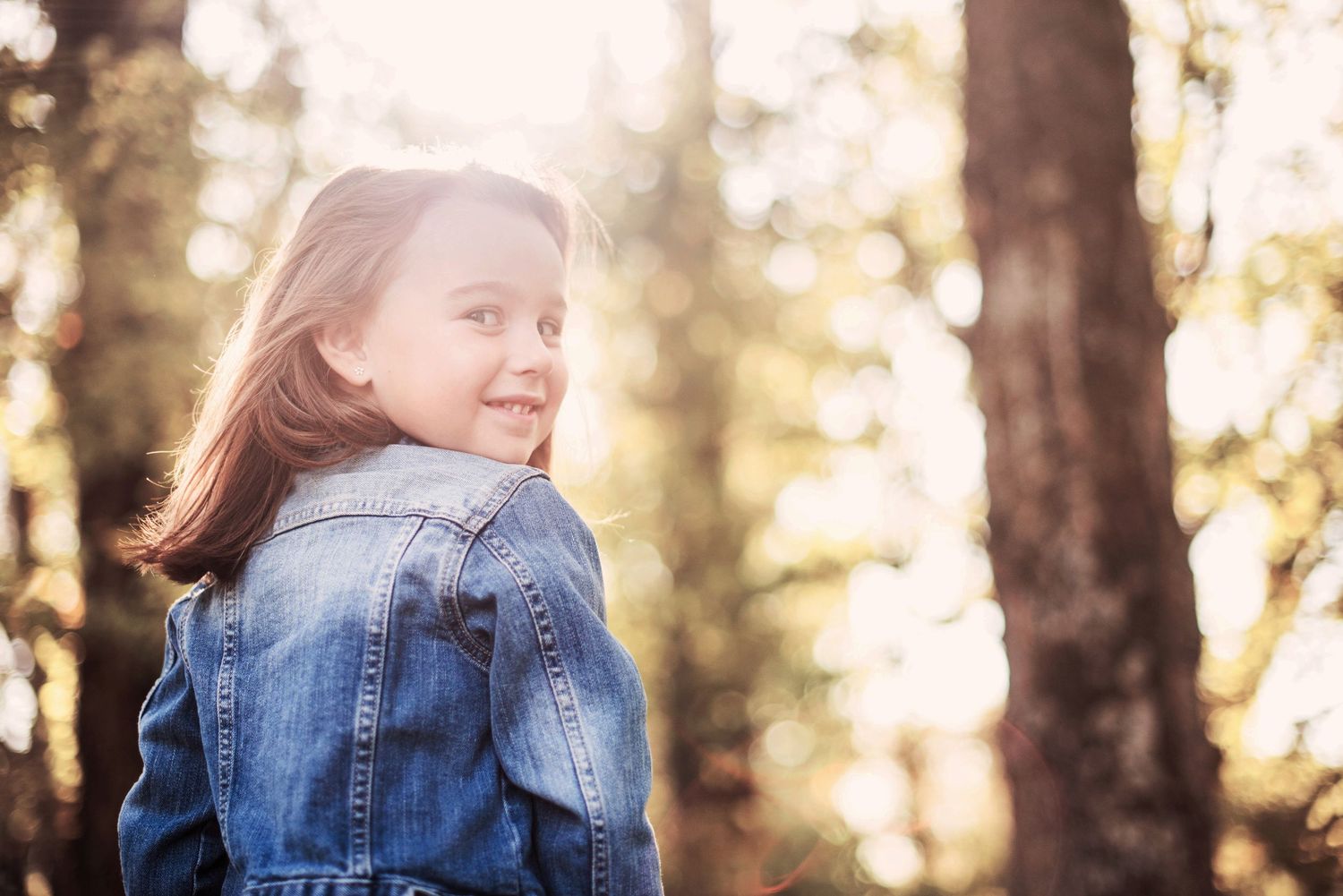 Child playing in sunlit forest wearing denim jacket.