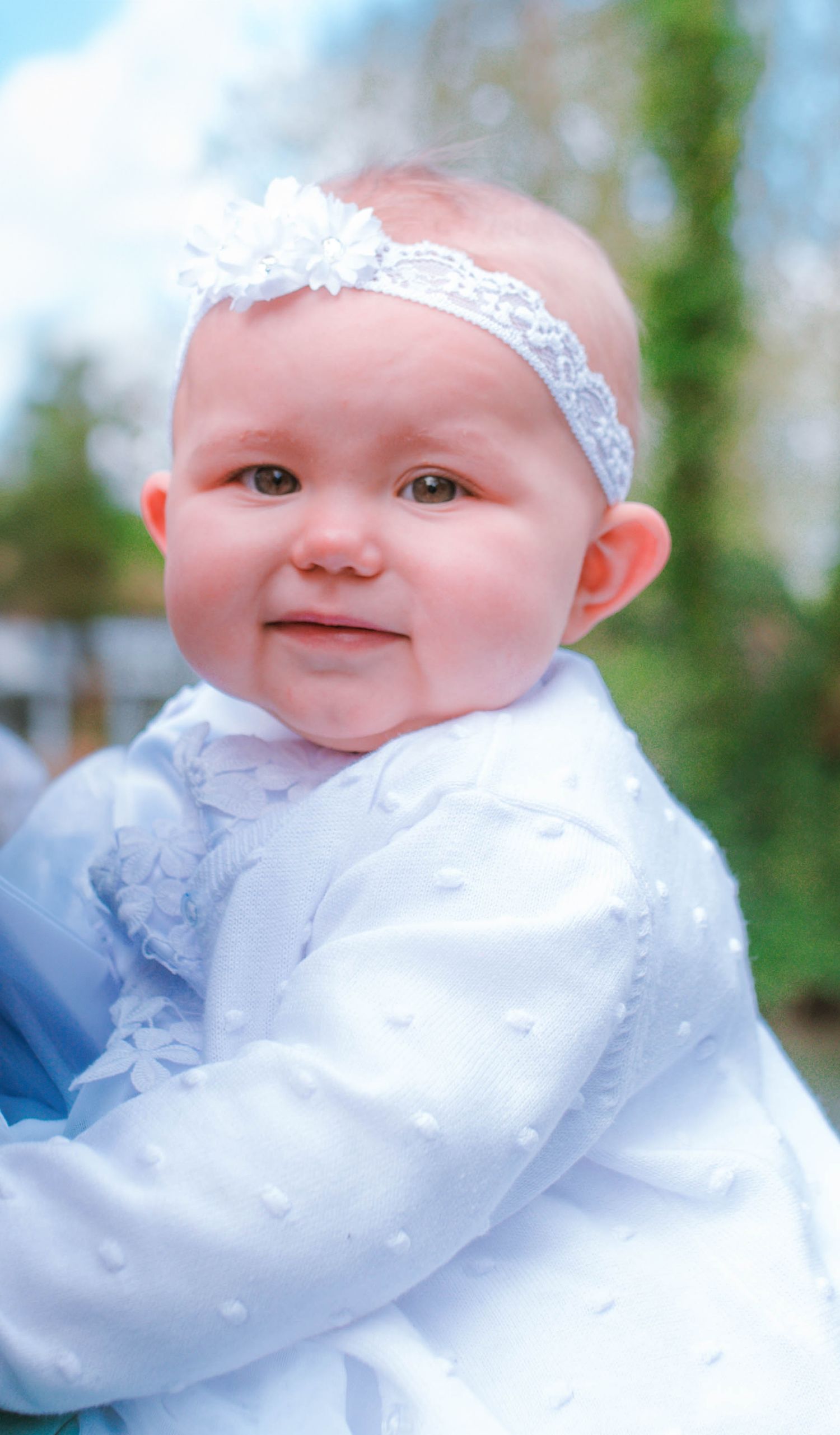 Baby wearing white lace dress and headband smiling at camera in outdoor setting.