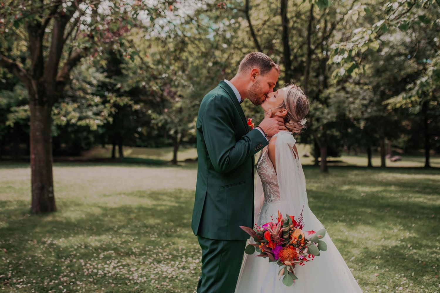 Romantic wedding couple poses in a sunlit garden with vibrant red and pink floral bouquet.