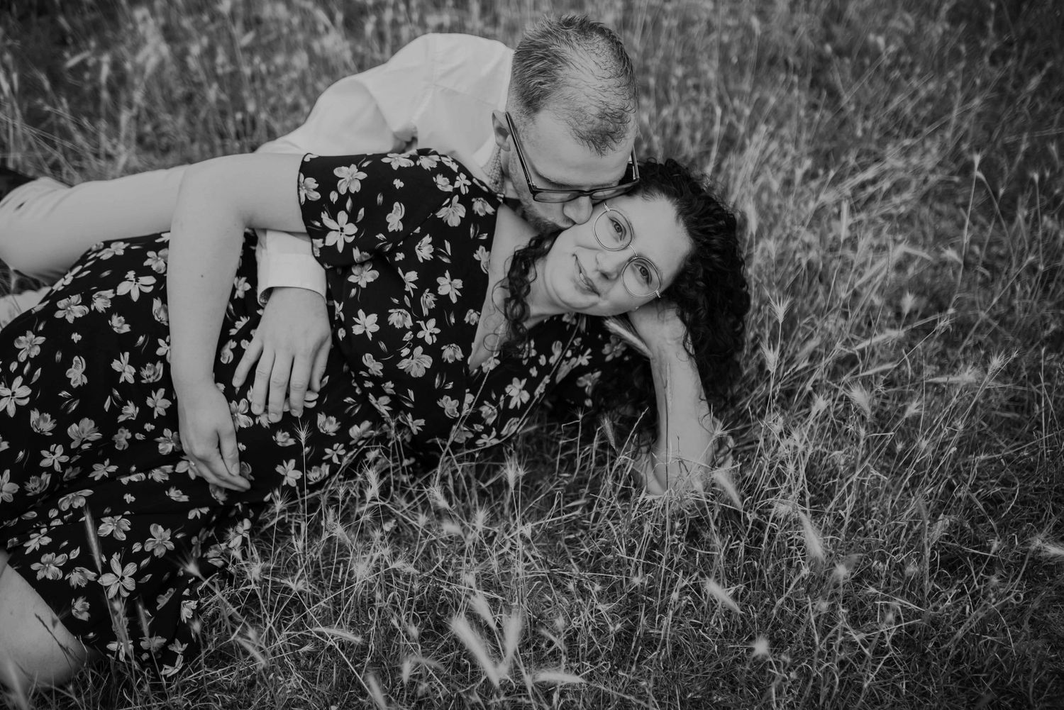 A black and white maternity photo captured in tall grass with soft natural lighting.