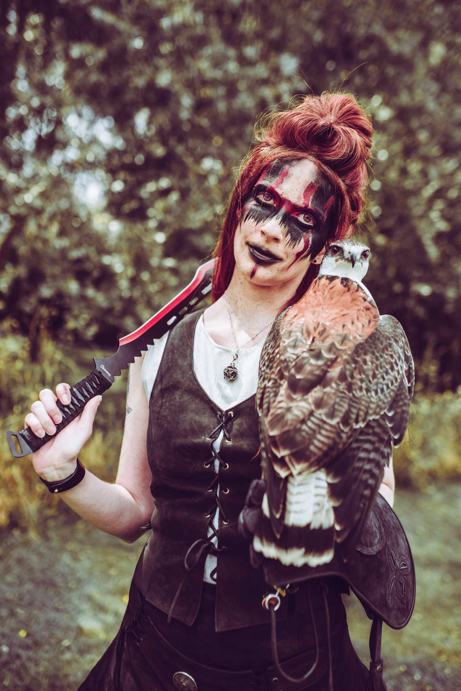 Gothic styled portrait with red hair holding a hawk in woodland setting.