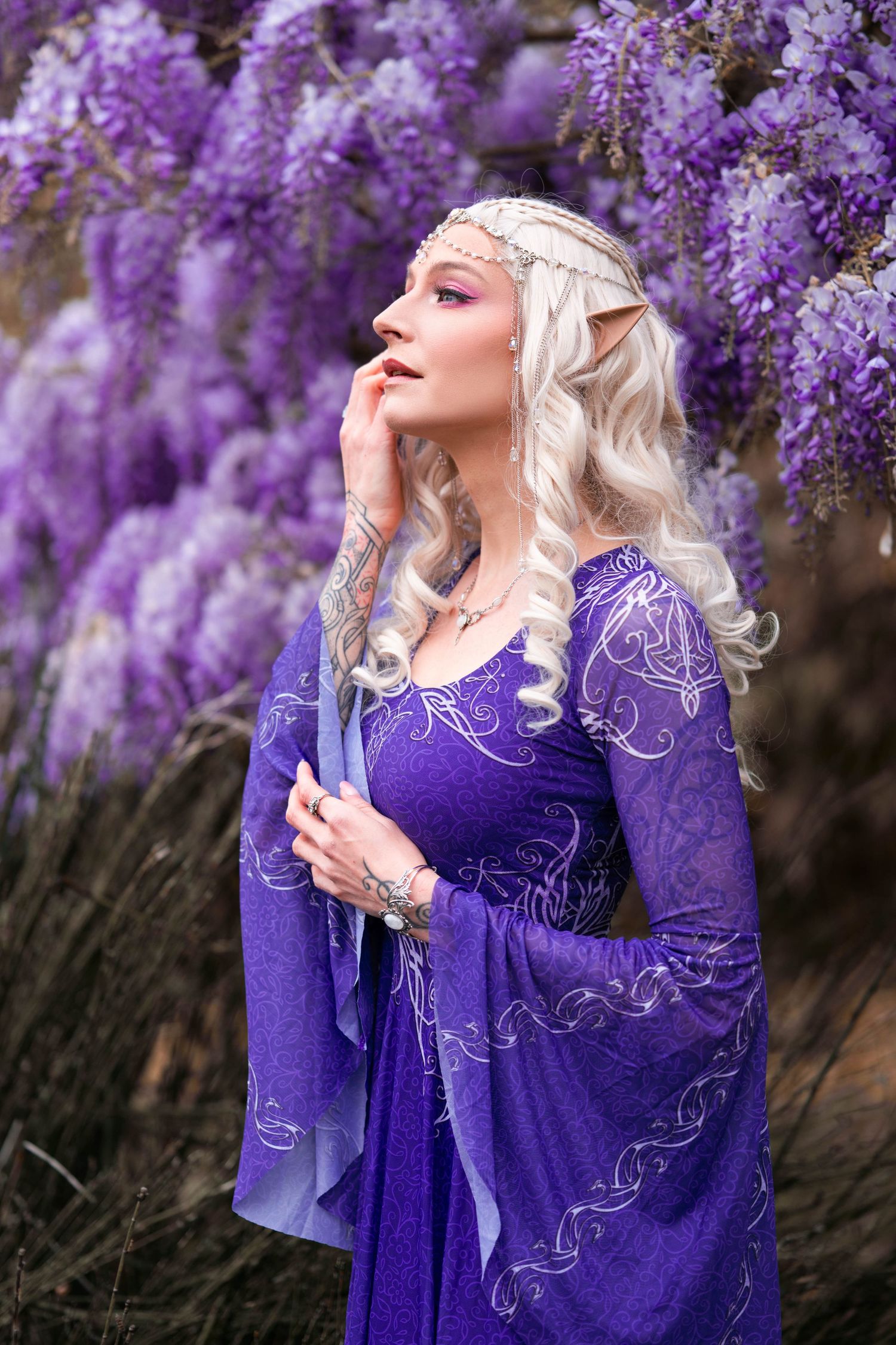 Blonde elf cosplayer in purple gown with pointed ears and silver crown poses before blooming wisteria flowers.