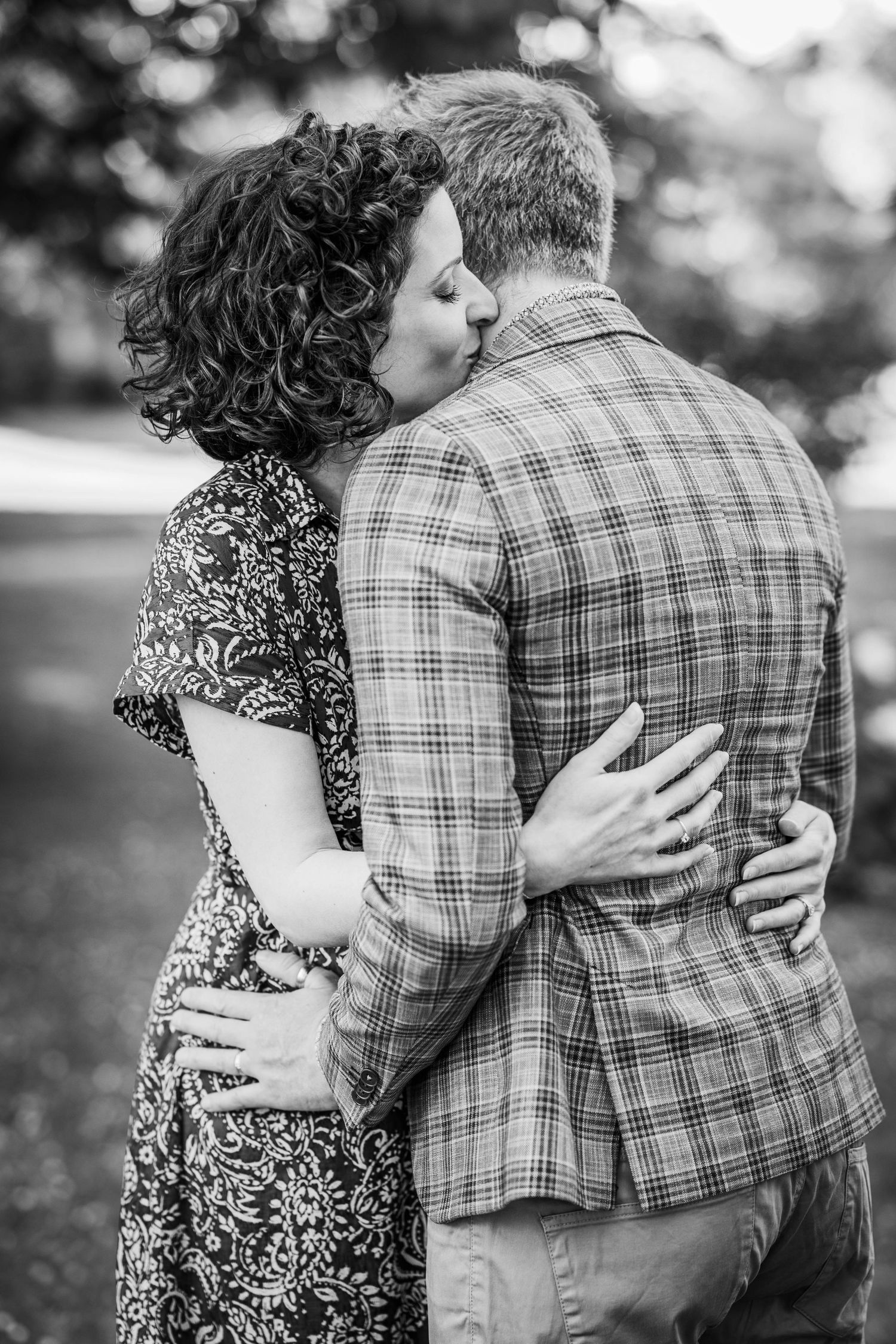 Black and white photo of a couple sharing a kiss outdoors, woman in floral dress embracing man in plaid blazer.