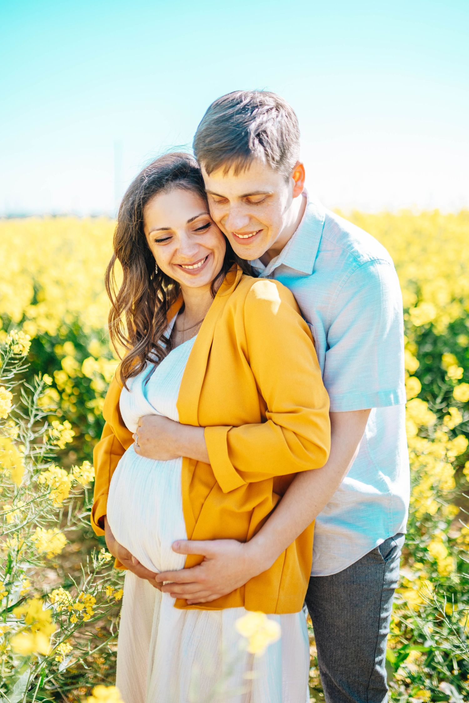 Happy pregnant couple embracing in a vibrant yellow flower field on a sunny day, woman in yellow jacket smiling.