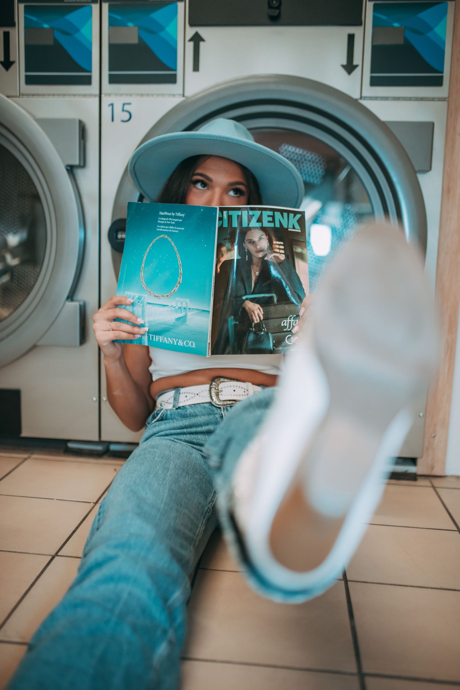 Creative laundromat photoshoot with turquoise hat and book against washing machines.