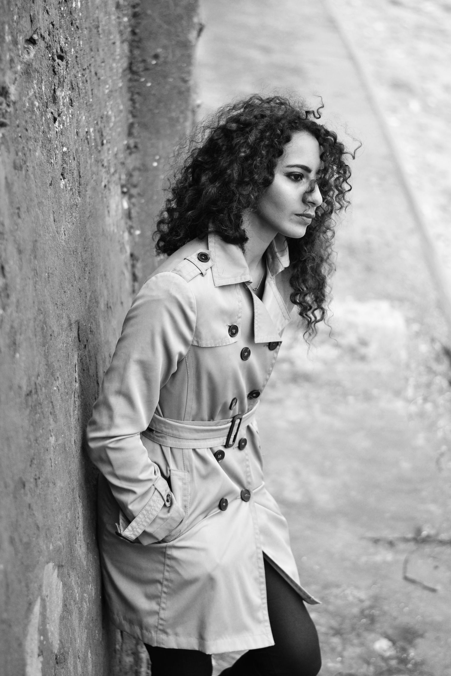 Model in beige trench coat poses dramatically against concrete wall.