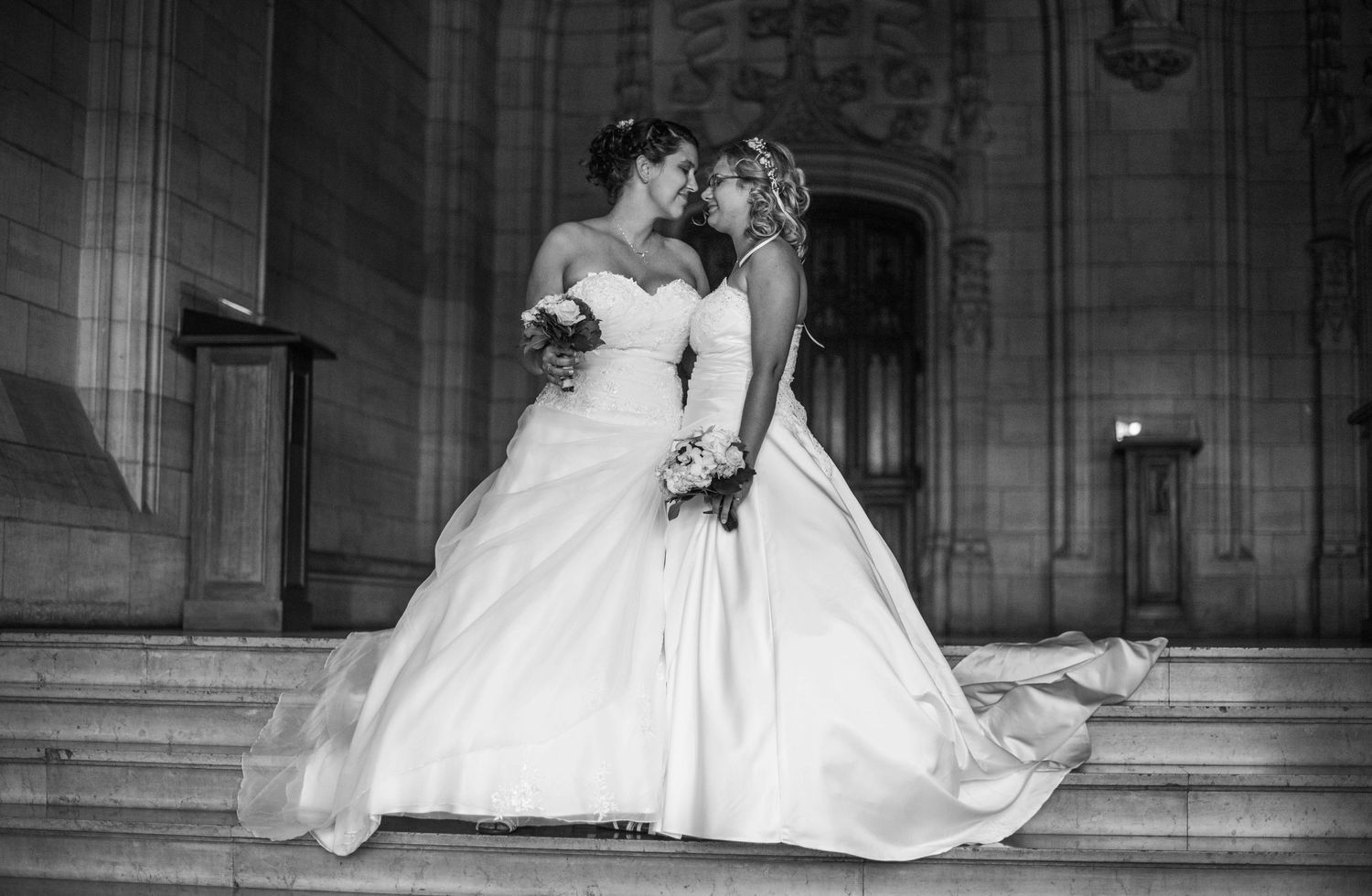 Two brides in elegant white gowns share a romantic moment on stone church steps.