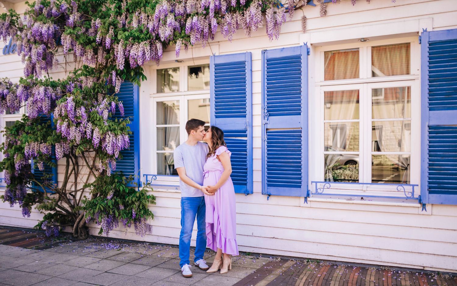 Couple kissing under purple wisteria blooms beside a white house with blue shutters.