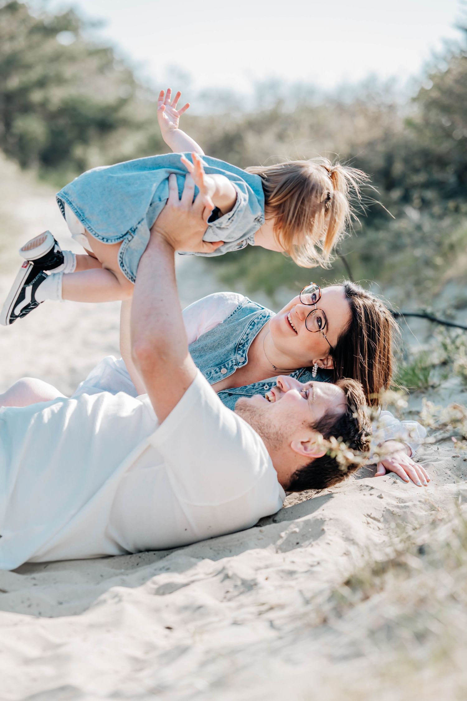 A family plays on sandy beach, father lying down lifting toddler while mother watches and smiles joyfully.