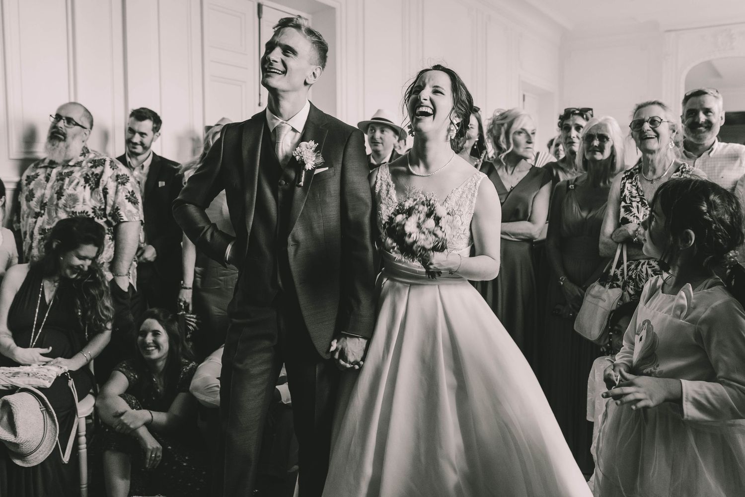 Black and white photo of a joyful bride and groom holding hands at their wedding ceremony surrounded by guests.