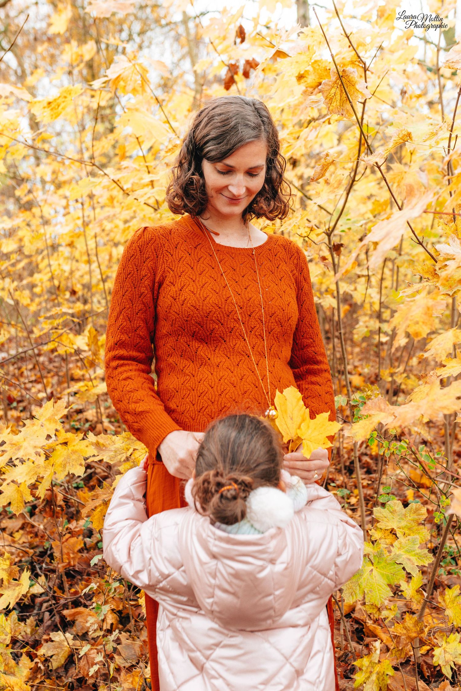 Mother in orange sweater plays with child against golden autumn leaves background.