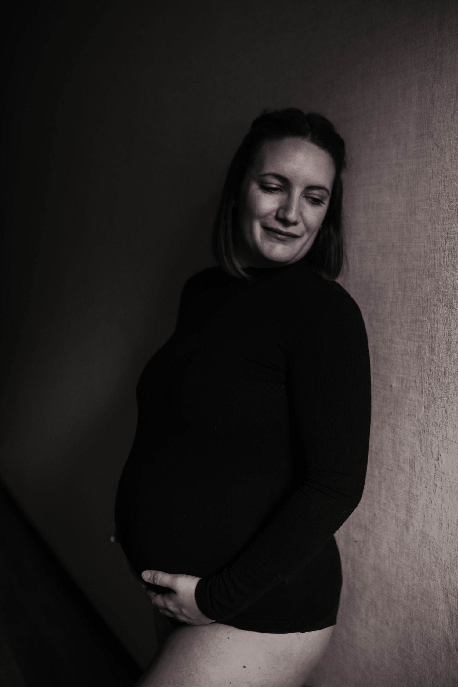 Black and white portrait of pregnant woman in dark sweater against wall.