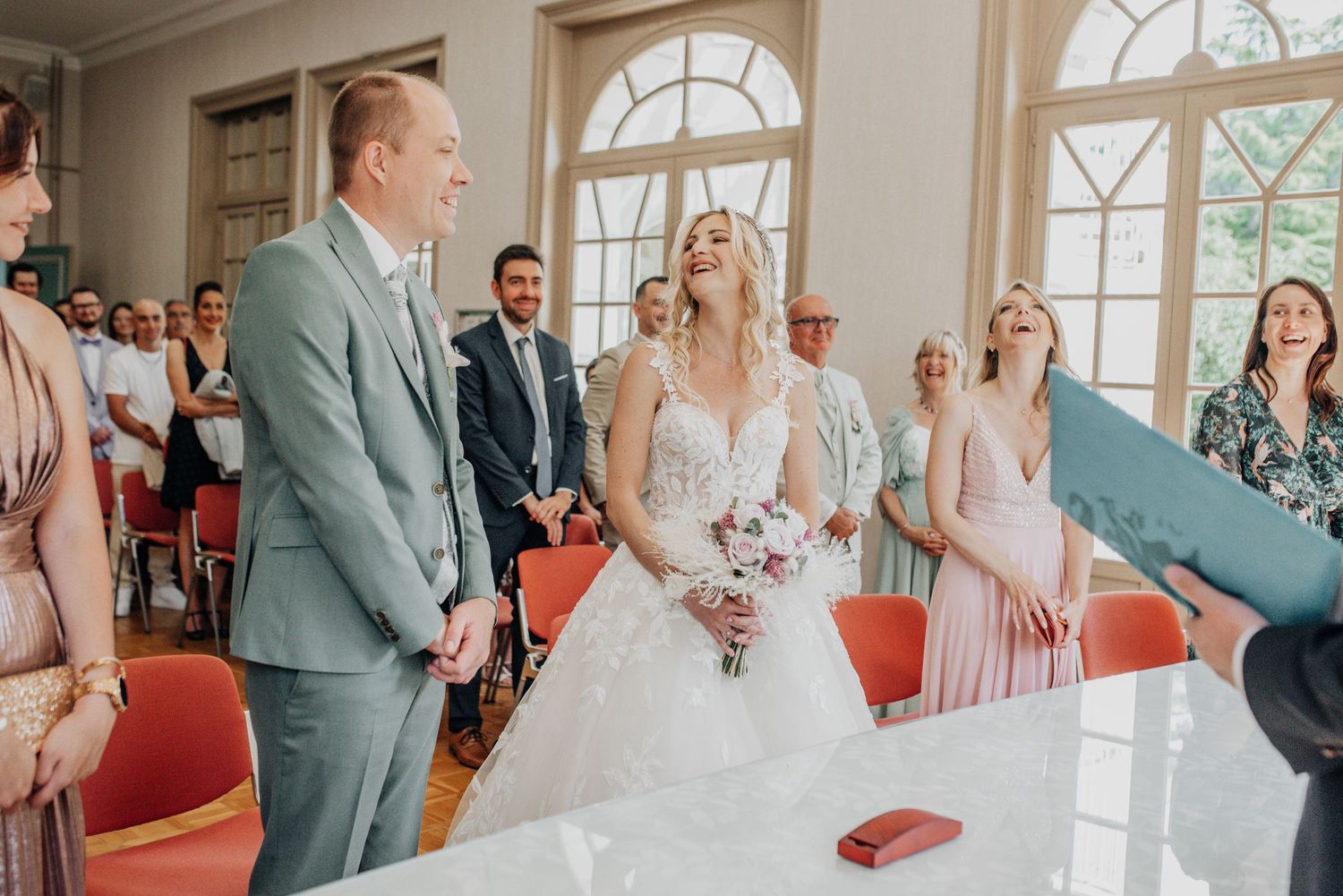 Intimate indoor wedding ceremony with guests in formal attire and coral-colored chairs.