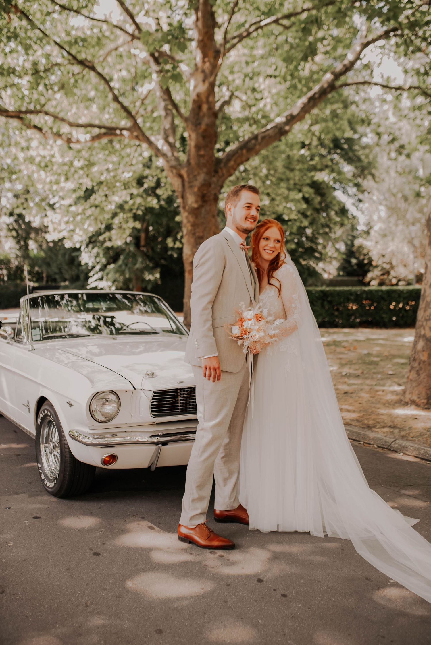 Wedding couple poses with a classic white vintage car under shady trees.