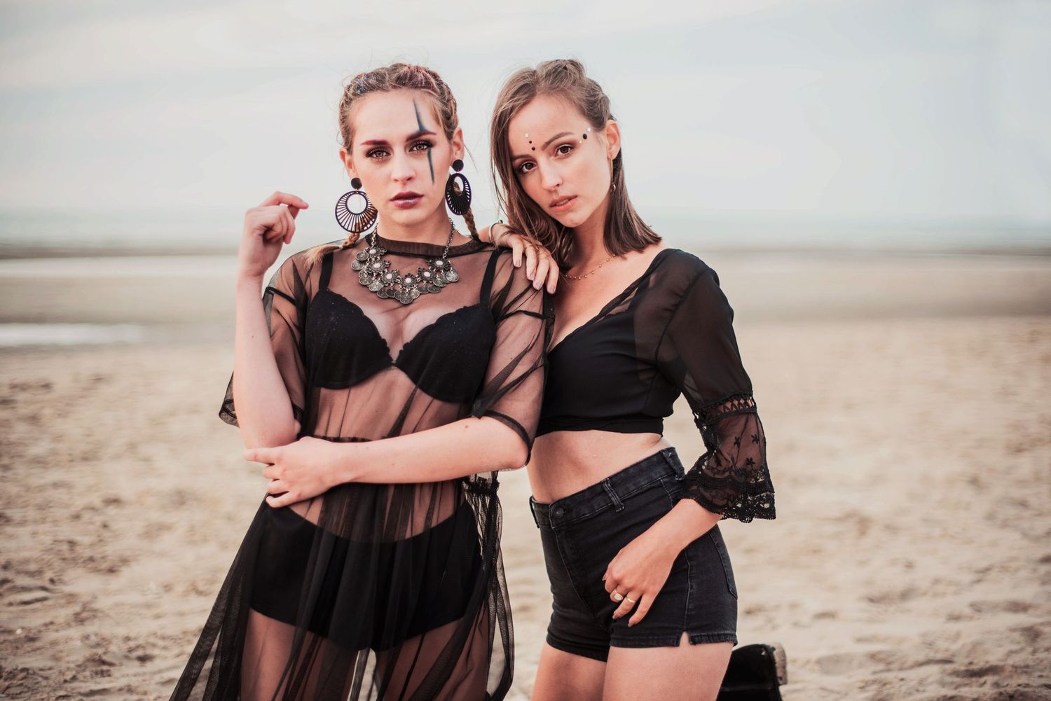 Two models in black outfits pose dramatically on a sandy beach during sunset.