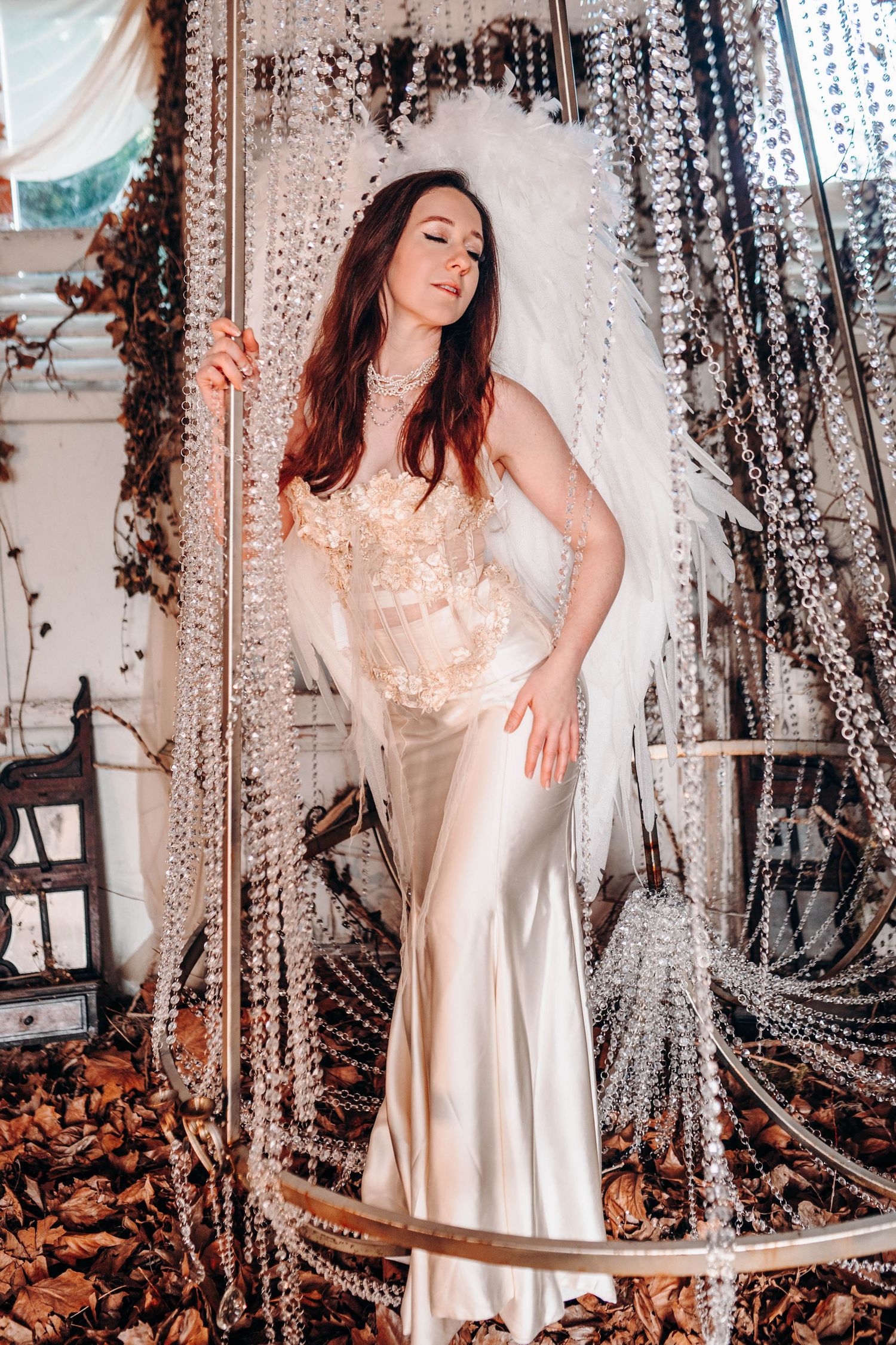 Woman in ivory satin gown with white feather wings poses among crystal bead curtains in elegant setting.