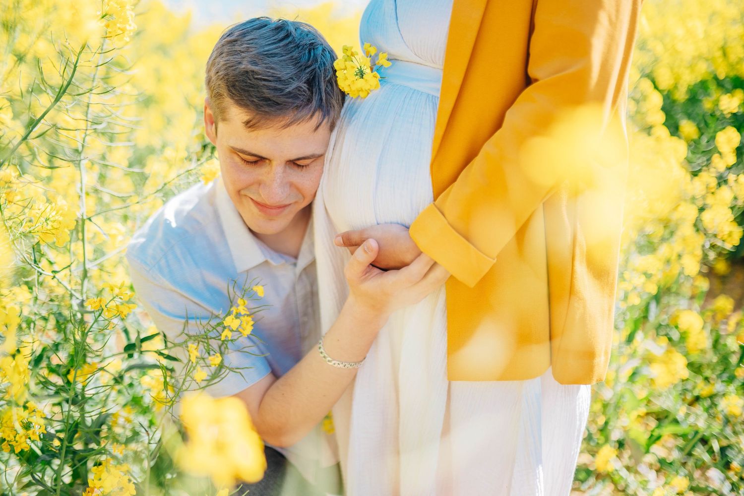 Man kneeling in yellow flower field listening to pregnant woman's belly, romantic maternity photo outdoors.