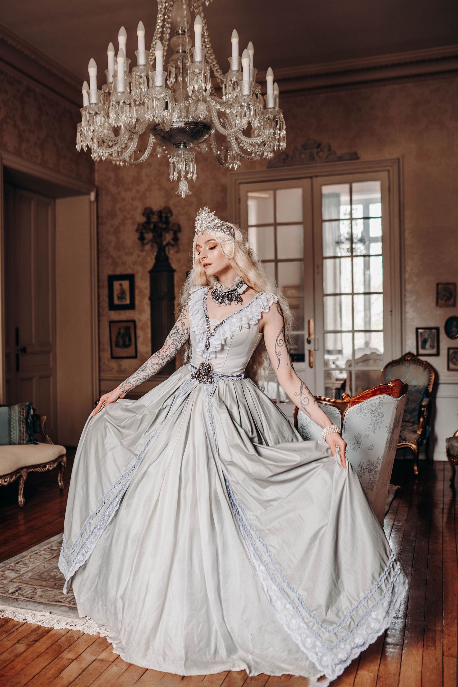 A blonde woman in an elegant silver ball gown and crown poses in a vintage room beneath a crystal chandelier.