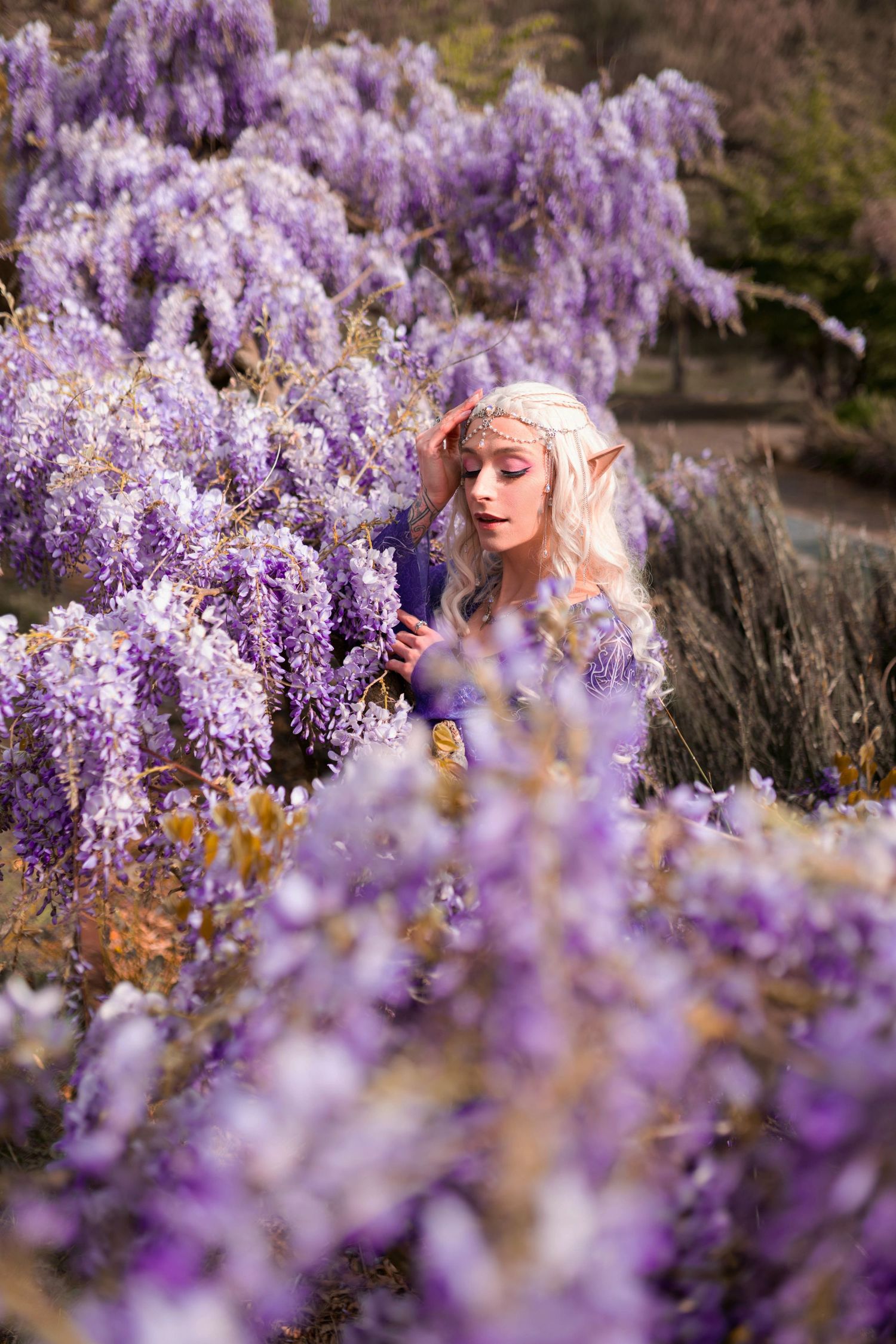 A blonde elf cosplay model with pointed ears poses among cascading purple wisteria flowers in an enchanting outdoor setting.