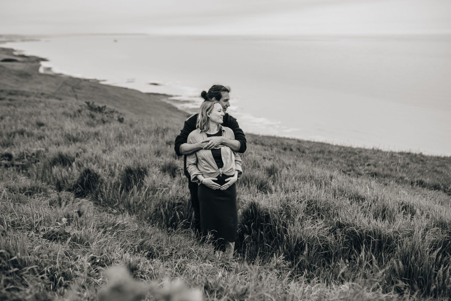 A series of intimate maternity photos taken on a grassy hillside against an overcast sky.