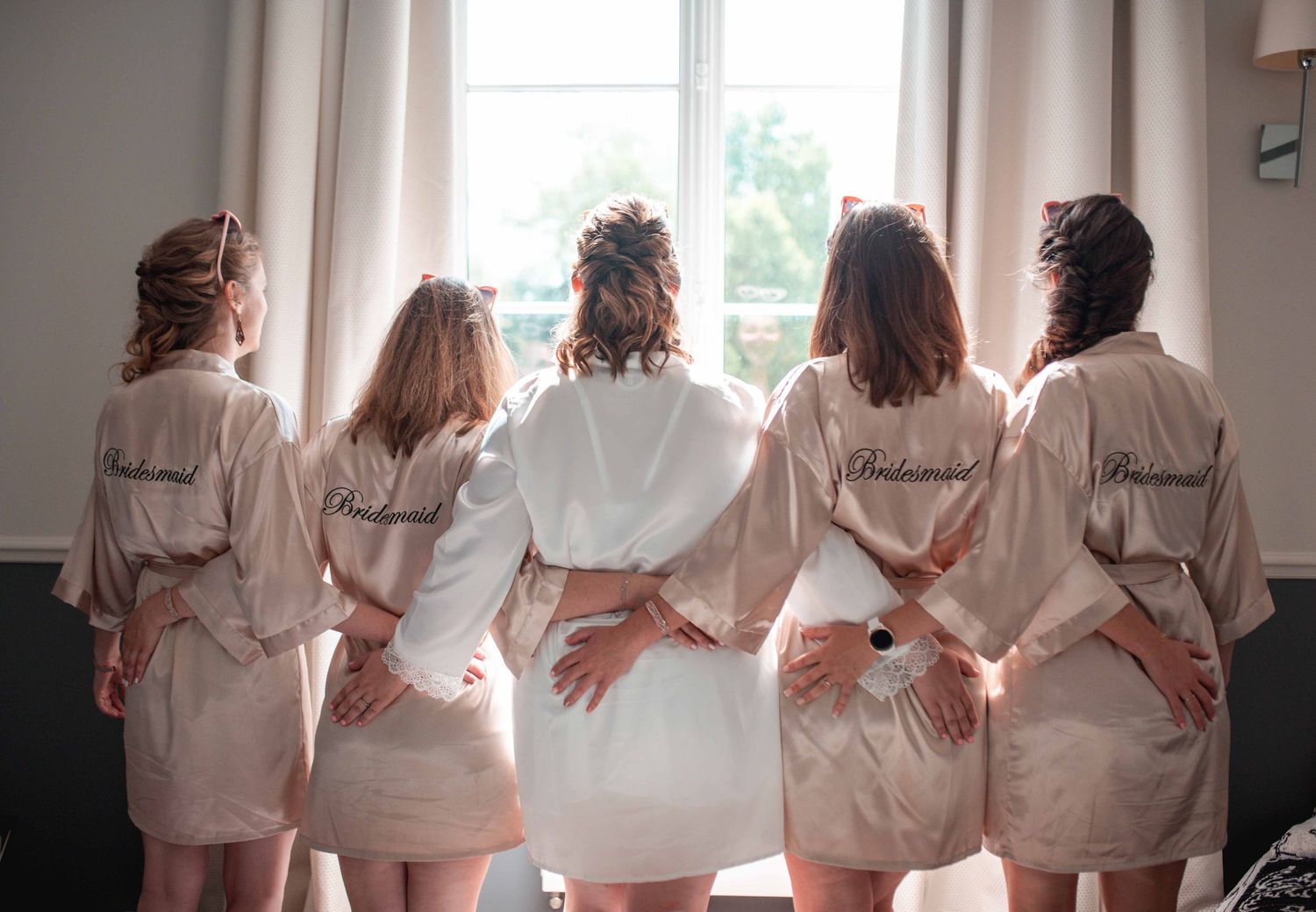Five bridesmaids in matching silk robes standing with their backs to the camera by a bright window.