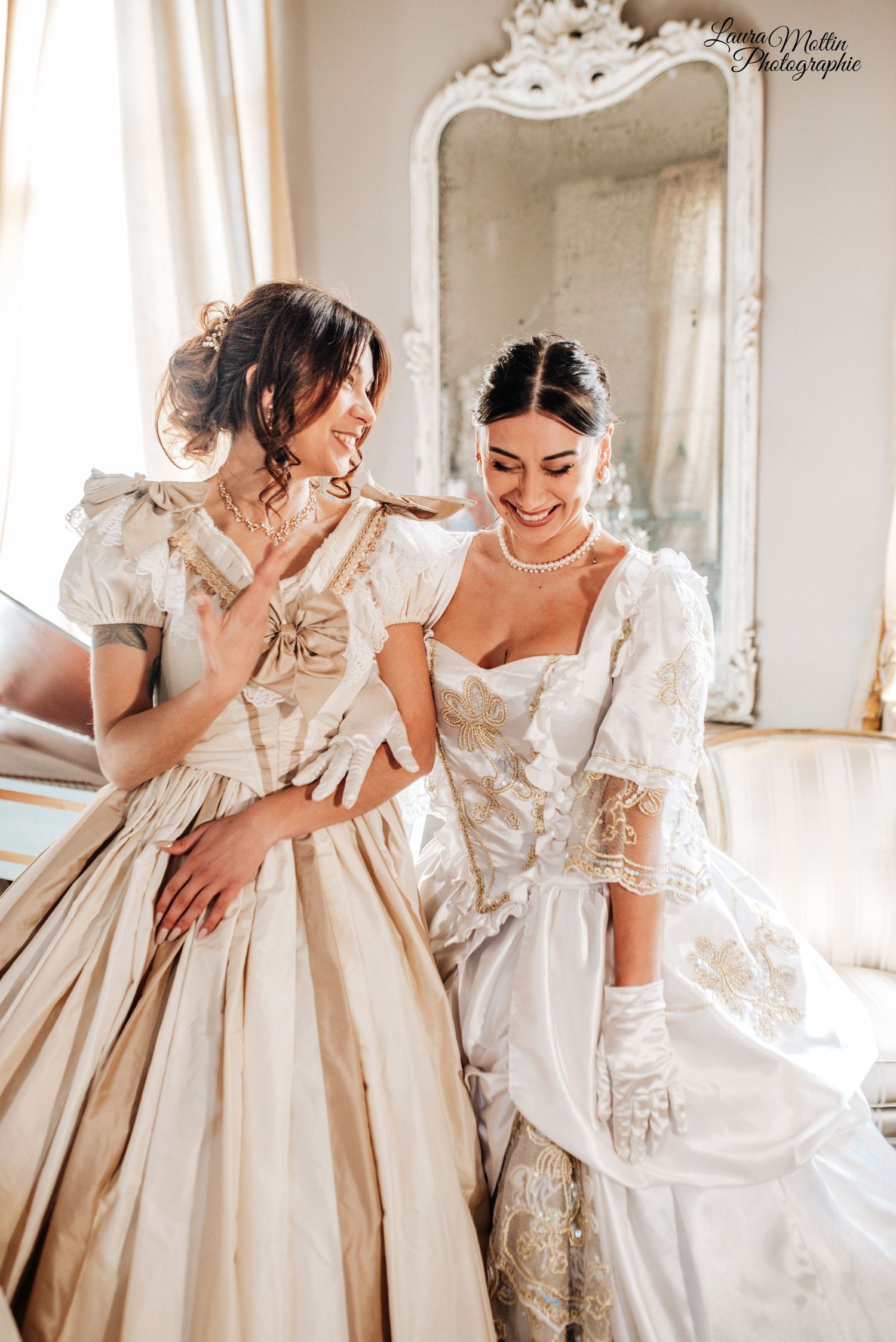Ladies in ornate period ball gowns sharing a joyful moment in a sunlit room.