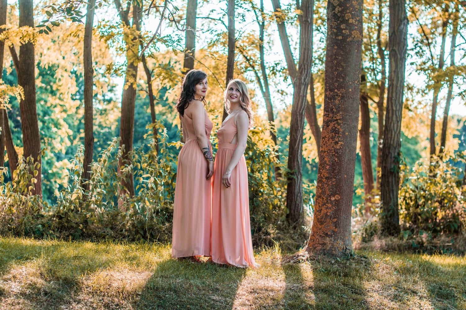 Row of pink bridesmaid dresses visible through sunlit forest trees.
