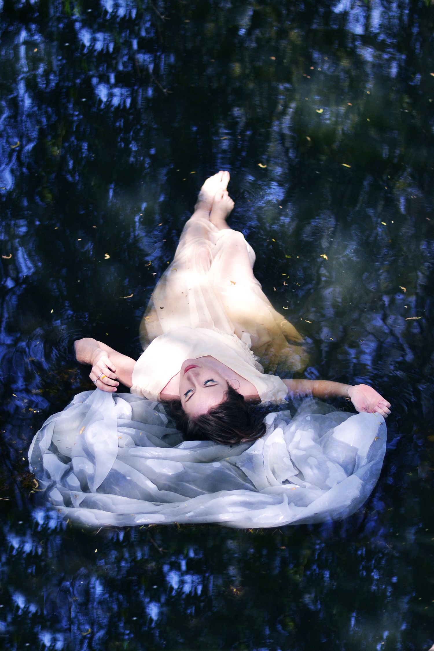 Ethereal figure in white flowing dress floating in dark water creating artistic underwater portrait.