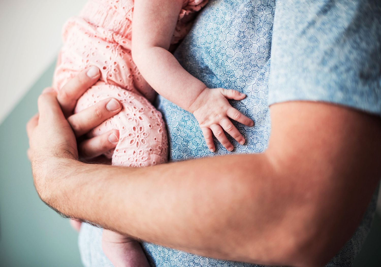 Gentle hands cradle sleeping newborn wrapped in pink swaddle against blue fabric.