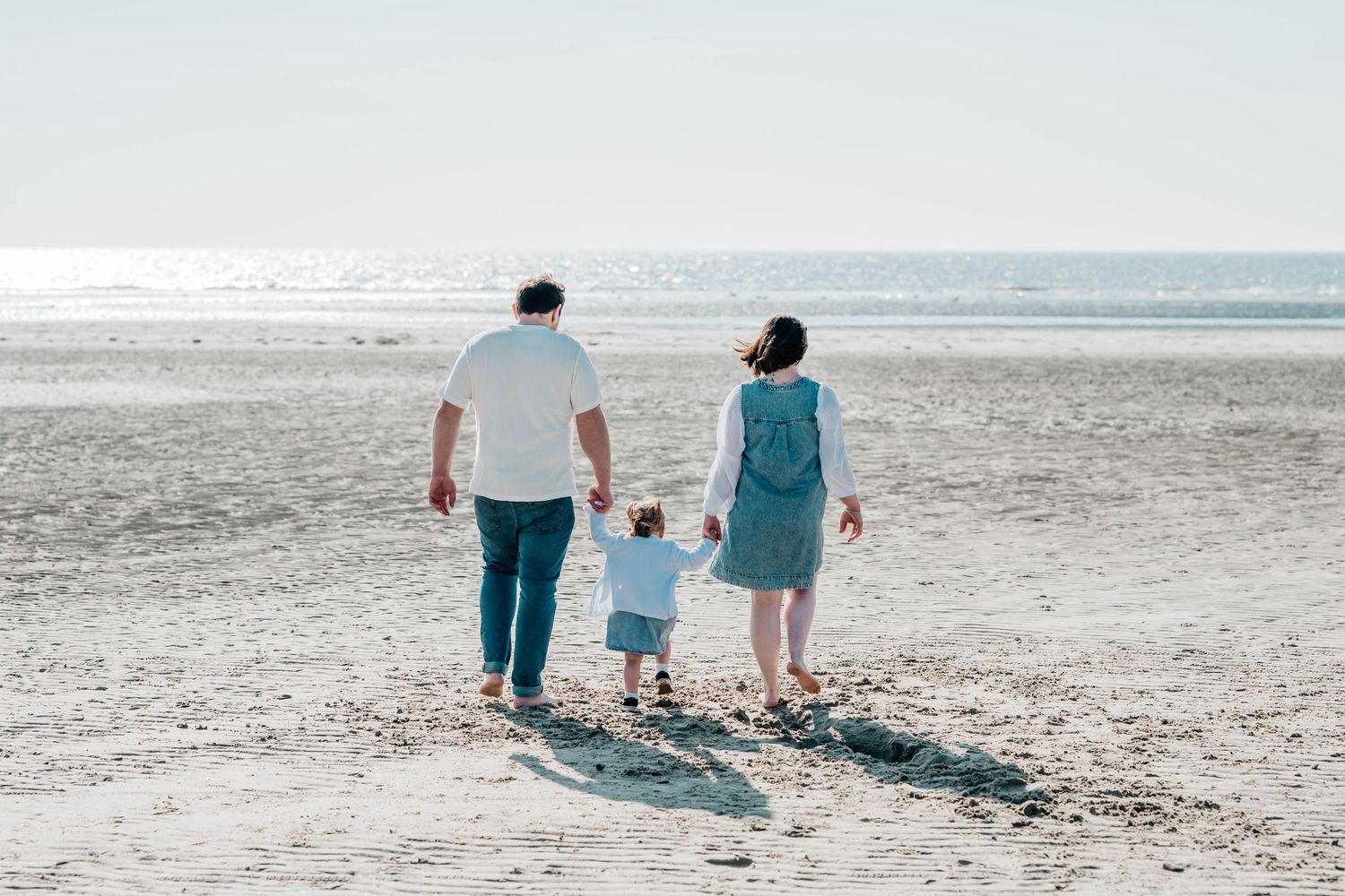 Family of three walking hand-in-hand on a sandy beach toward the calm ocean on a bright, hazy day.