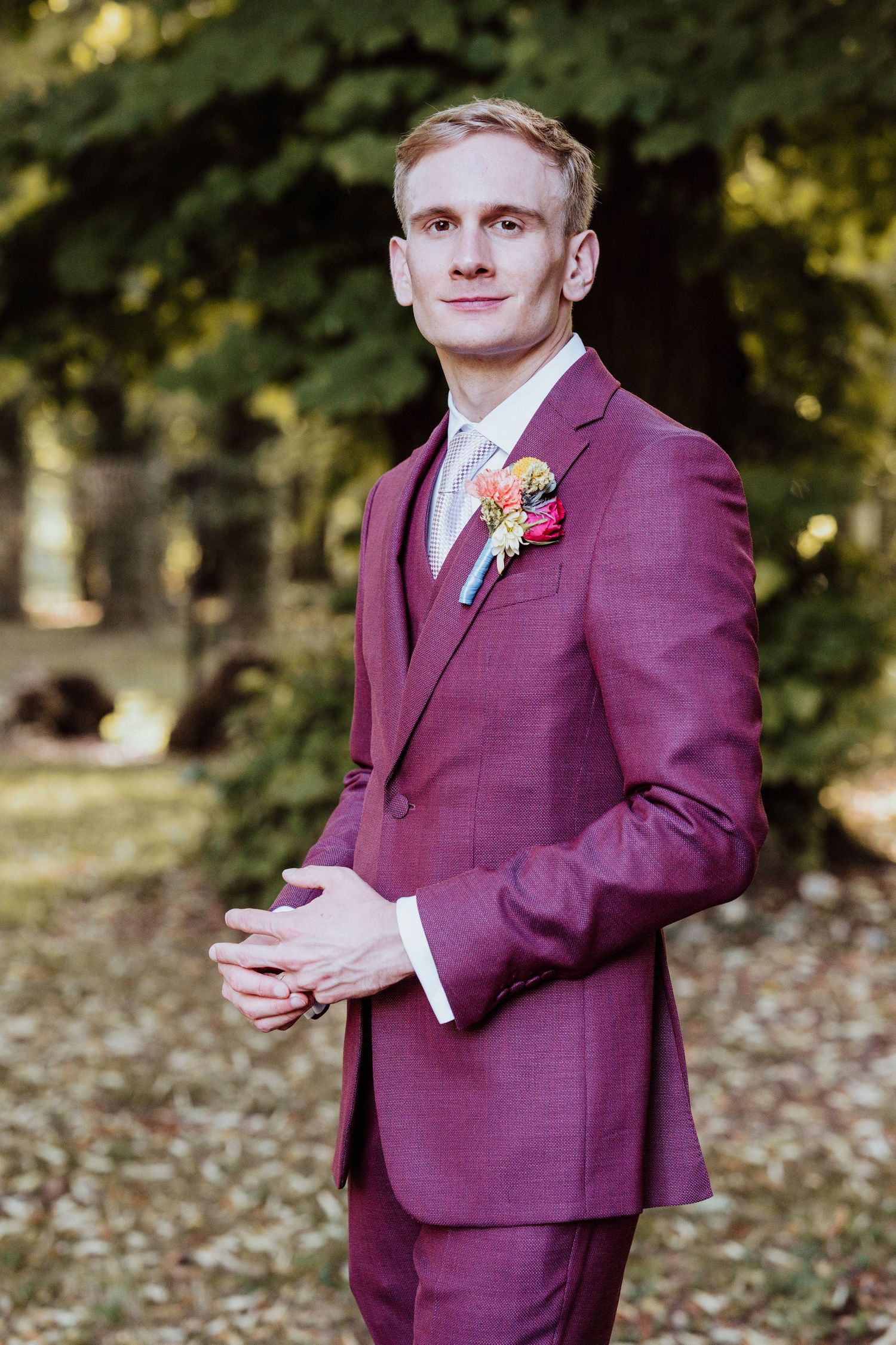 Young groom in burgundy three-piece suit with floral boutonniere poses outdoors among autumn trees.