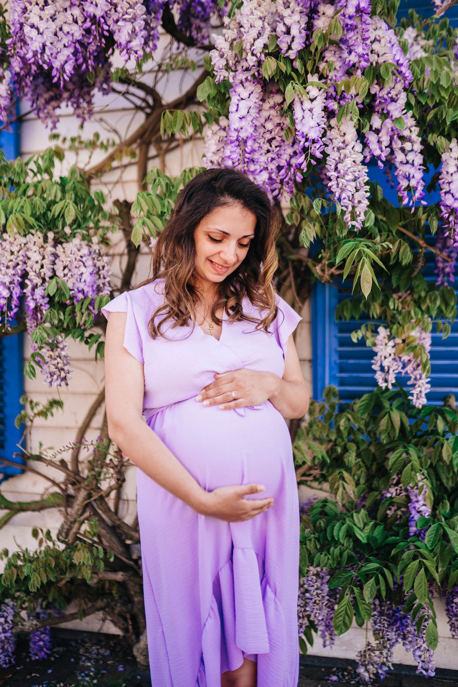 Pregnant woman in lavender dress cradling baby bump under blooming wisteria flowers outdoors.