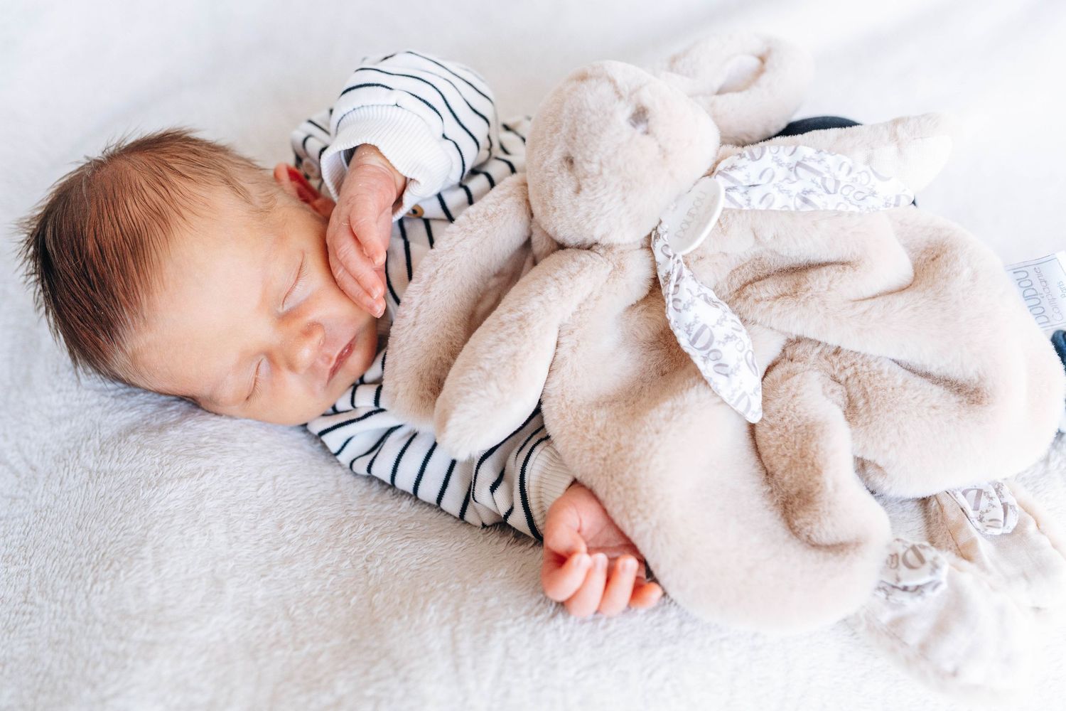 Newborn baby sleeping peacefully in striped onesie cuddling a soft plush bunny toy on a white blanket.