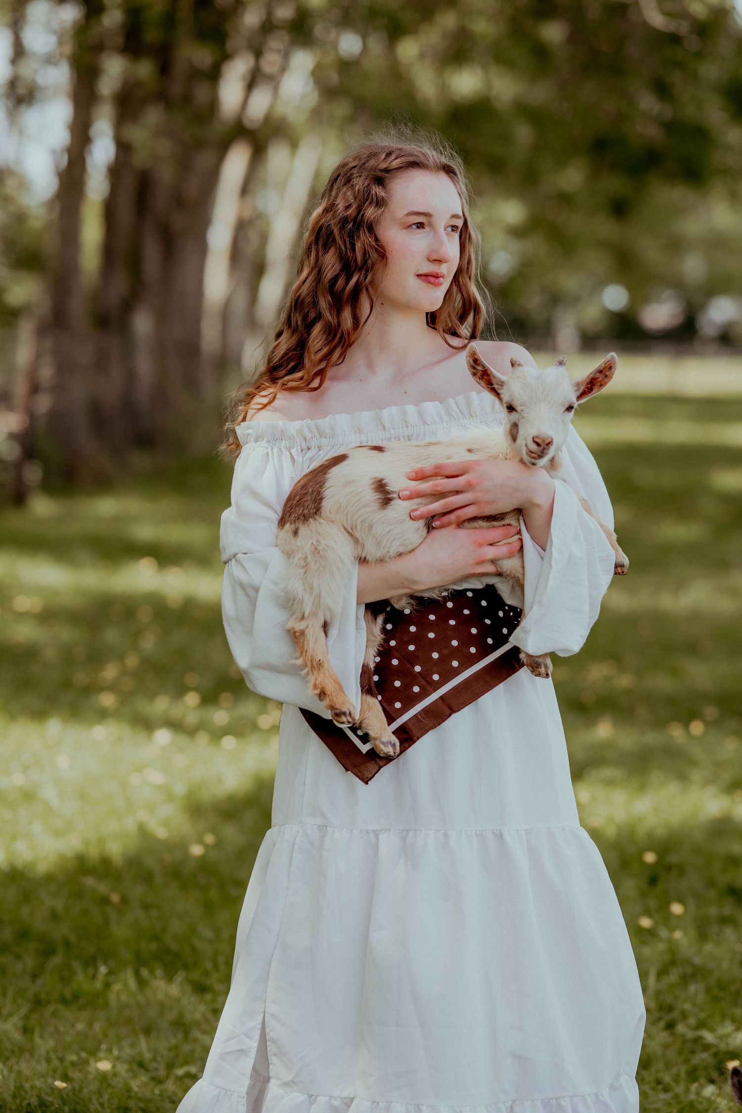 Young woman in white off-shoulder dress holding a baby goat outdoors in a green park setting.